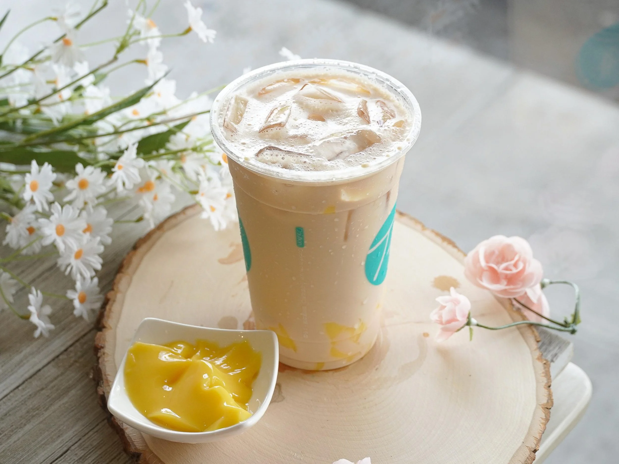A plastic cup filled with iced coffee or latte on a wooden surface with a small bowl of mango pudding or custard nearby. There are white flowers and a pink flower on the surface, with a white wooden tray in the background.