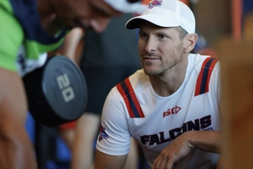 A man in a white sports jersey with red and blue accents, wearing a white cap, is sitting indoors, listening attentively to a person in a green shirt holding a dumbbell.