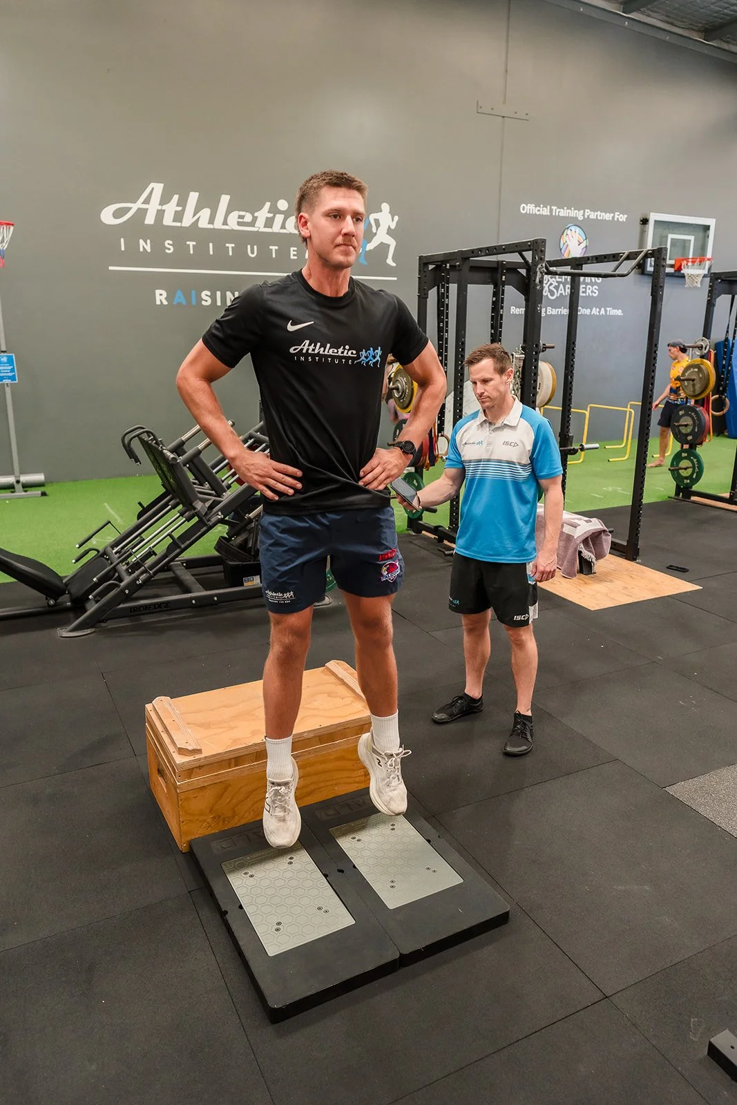 A man in athletic clothes stands on a measuring device in a gym with his hands on his hips, while another man checks his height with a measuring tape. There is gym equipment in the background.