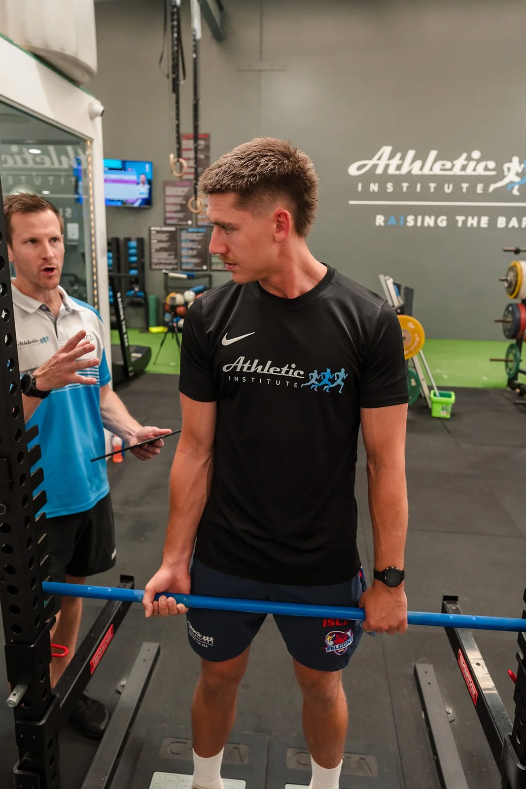 A young man with a muscular build lifts a barbell in a gymnasium while a coach or trainer speaks to him. The gym has a sign on the wall that reads 'Athletic Institute' and 'Raising the Bar'. The young man is wearing a black athletic shirt with the logo and text, navy shorts with a logo, and a watch. The trainer is wearing a blue and white shirt with the same logo as the young man.