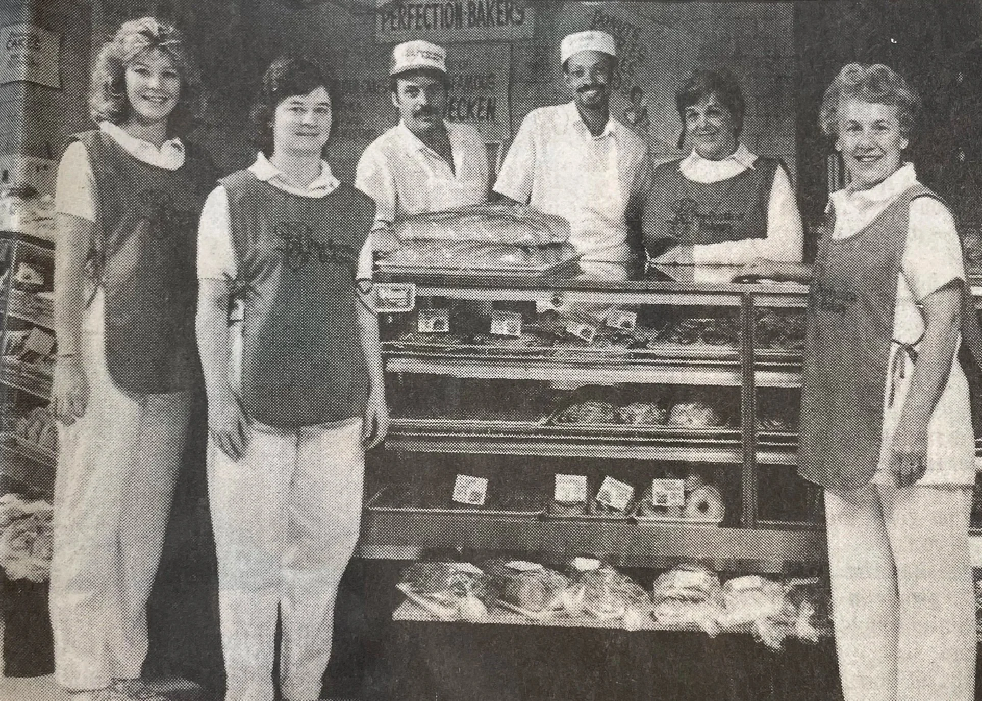 Black and white image depicting four bakery staff, standing around a full bakery case. There is a woman in front of the case, and three people behind the case. Hurtis is at the center, wearing a chefs apron and white chef hat. 
