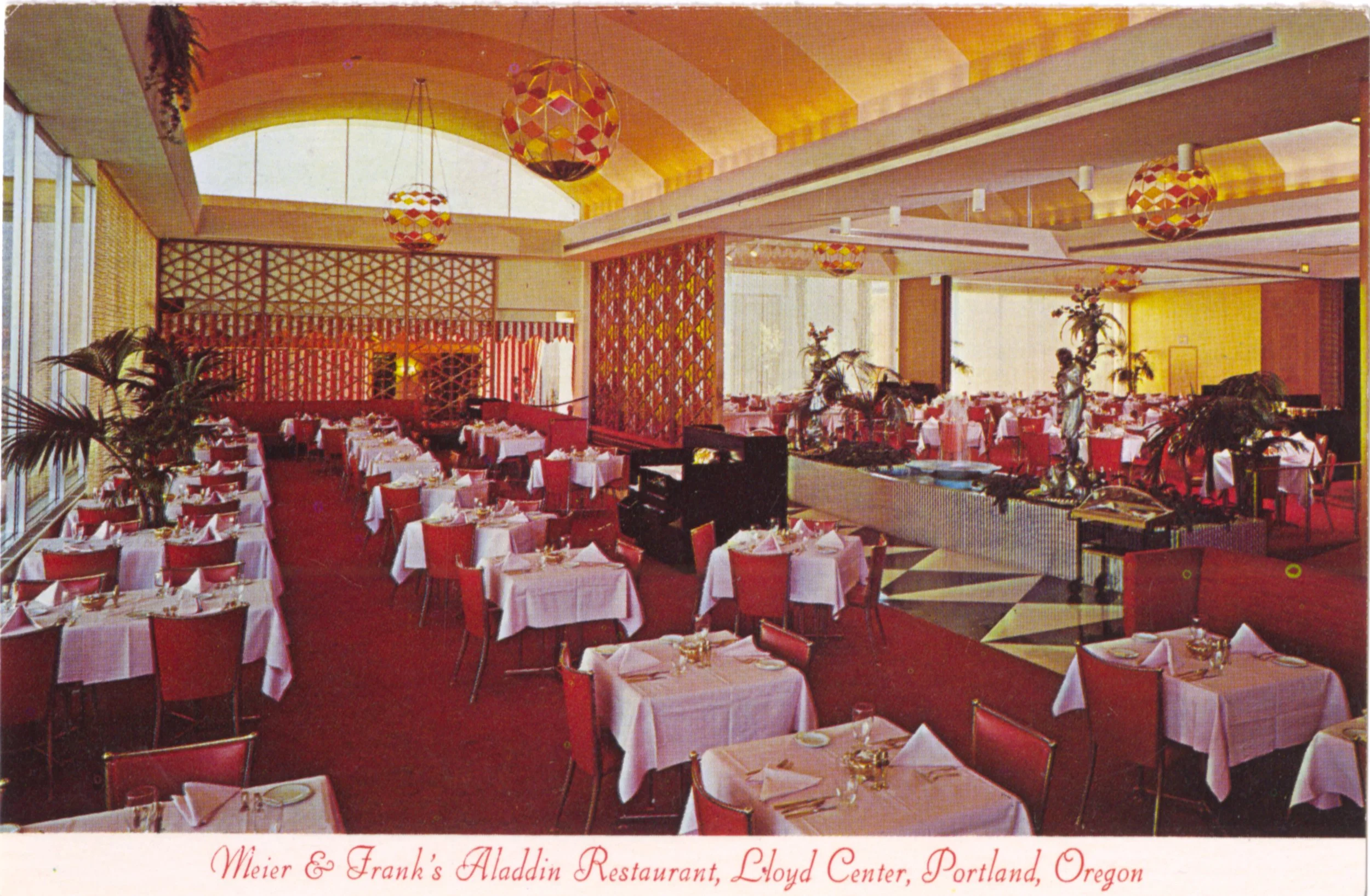 Postcard depicting interior of Meier and Frank's Aladdin restaurant. Red carpets and chairs, and white tablecloths on tables. Yellow and white striped ceilings and colorful chandeliers.