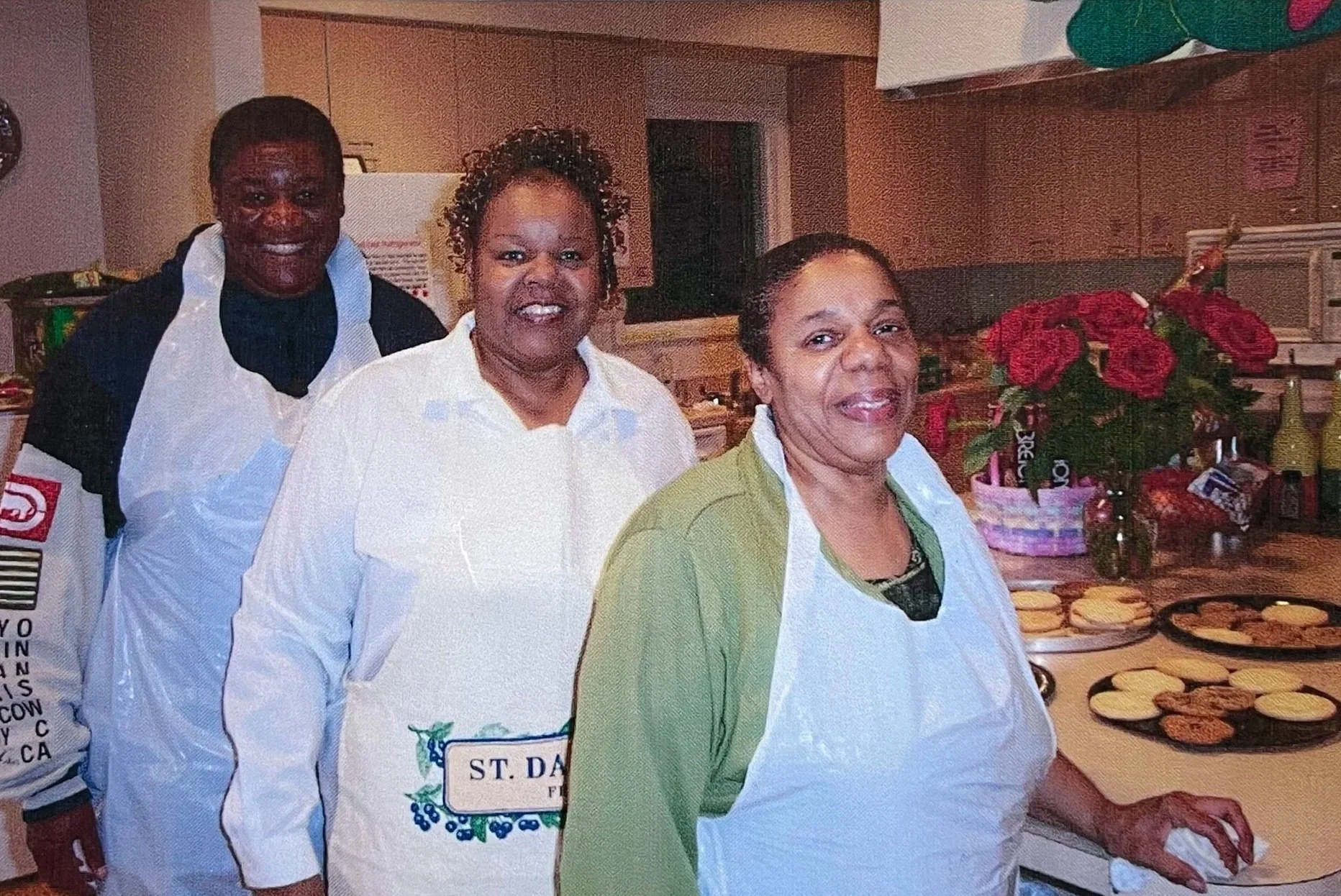 Dorothy standing between a woman and a man. All three are inside a kitchen, wearing plastic aprons. Dorothy is wearing a white top, the woman is wearing a green top, and the man is wearing a black top. All are smiling. 