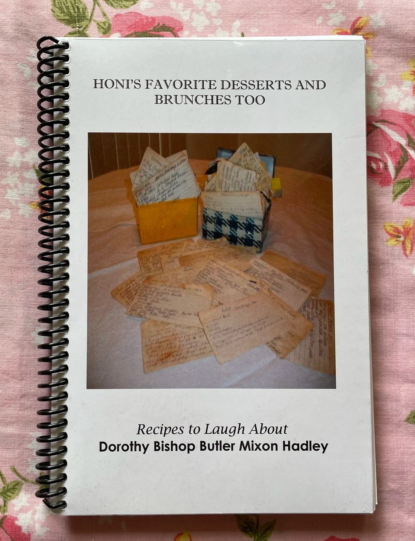 Cookbook sitting on top of pink sheets. Cover of cookbook is white with a photograph of hand-written recipe notecards that have yellowed with age. 
