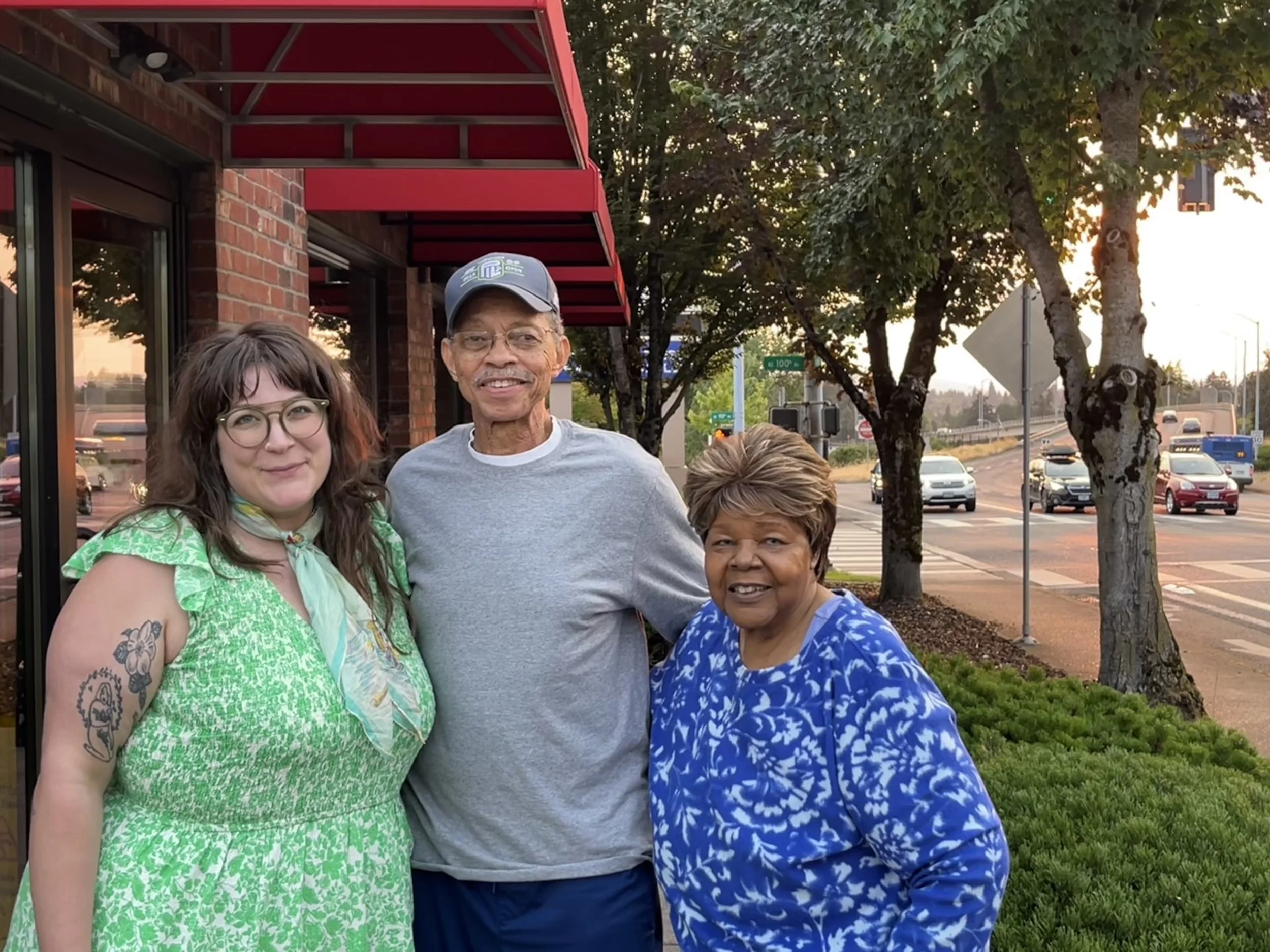 Sarah Harris, Hurtis Hadley, and Dorothy Hadley standing together outside of a restaurant. All three are smiling. Sarah is wearing a green dress, Hurtis is wearing a grey top, and Dorothy is wearing a blue top. 
