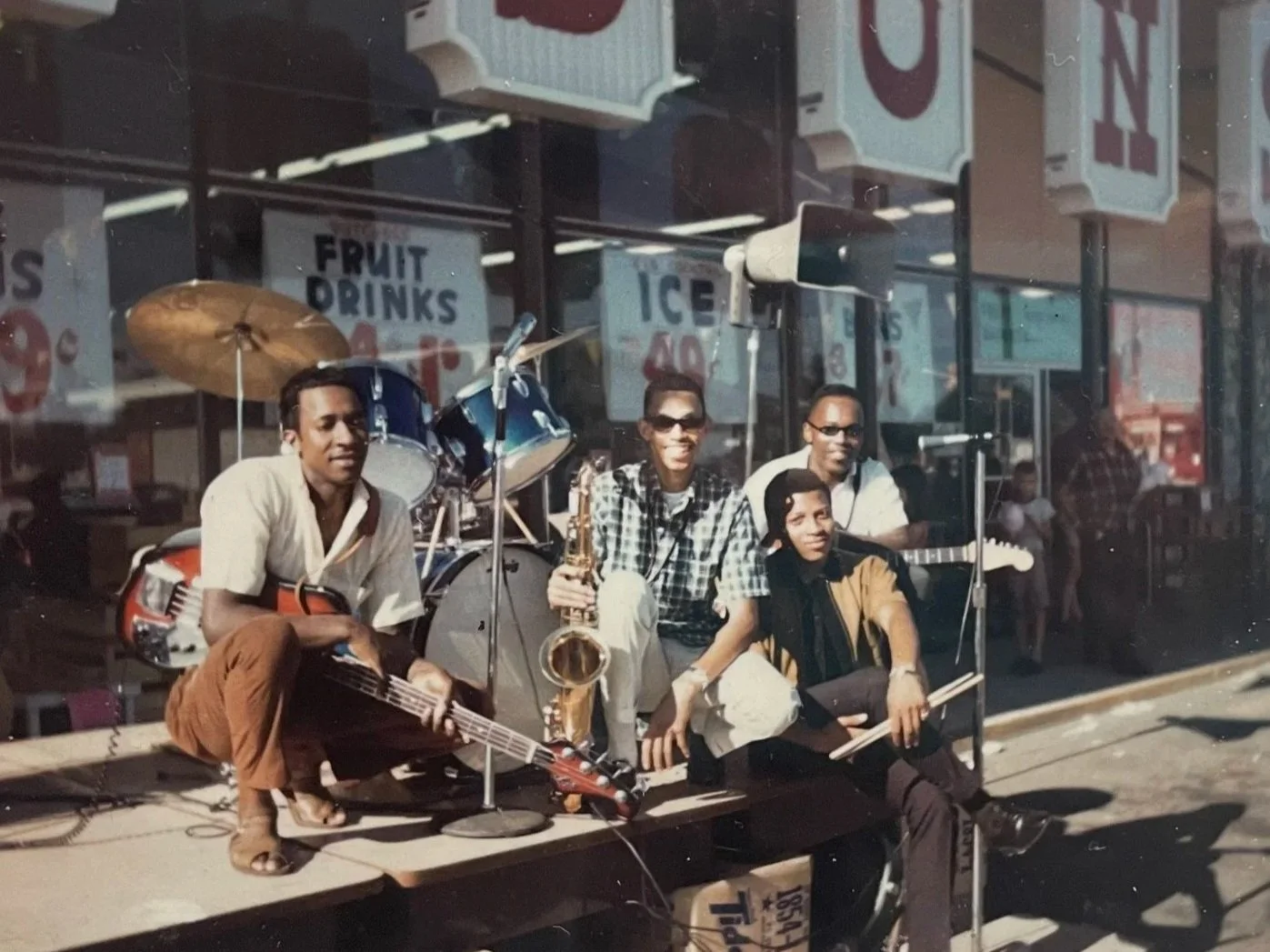 Hurtis and his band performing outside of Albertsons. Hurtis is hunched over holding a saxophone. He is surrounded by bandmates holding guitars and drum sticks. Albertsons signs in the background. 