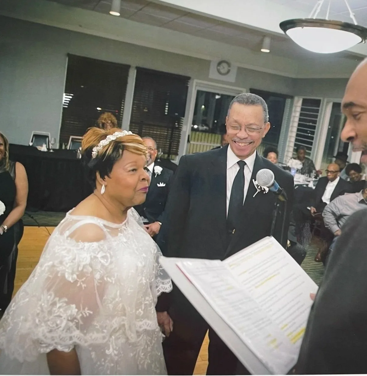 Dorothy Hadley wearing a white lace wedding dress and matching hairband. She is standing alongside Hurtis Hadley, who is wearing a black tuxedo and tie. Both are smiling and facing a pastor with an open book with notes. 