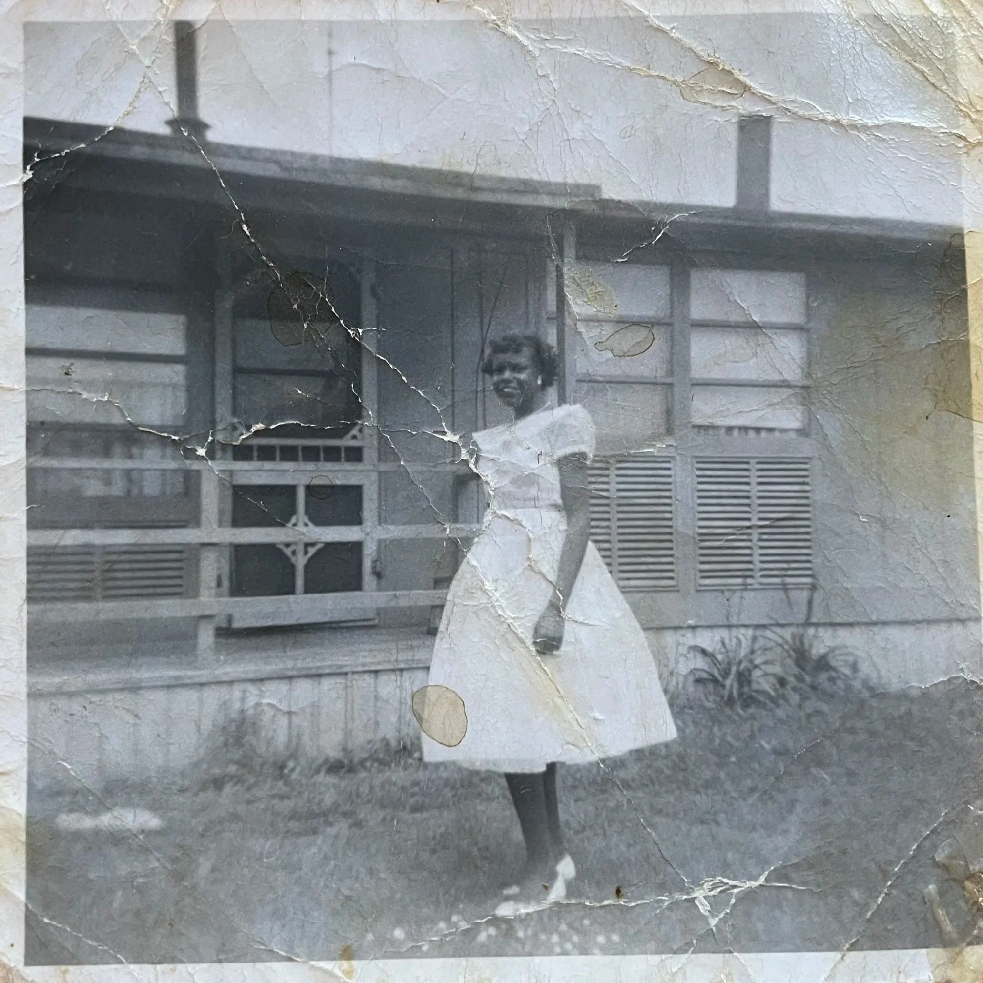 Dorothy standing in front of her house. She is wearing a white dress and underskirt, and white shoes. She is smiling and facing slightyl away from camera. The photograph itself is stained and bent. 