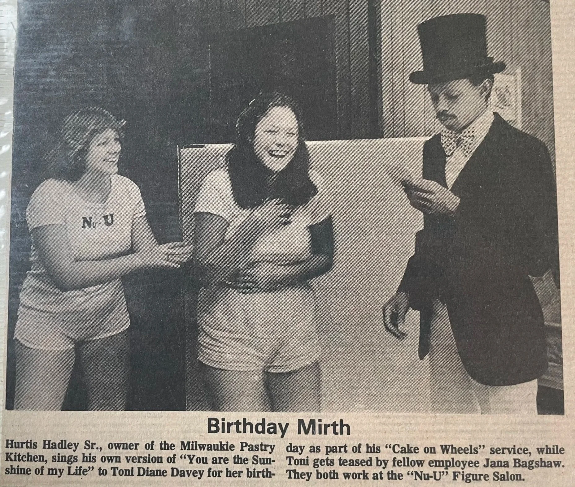 Black and white image depicting two girls laughing while Hurtis Hadley reads from a card. He is wearing a top hat and a alrge bowtie, and the girls are wearing white tops and shorts. "Birthday Mirth" is printed in black ink under the photo.