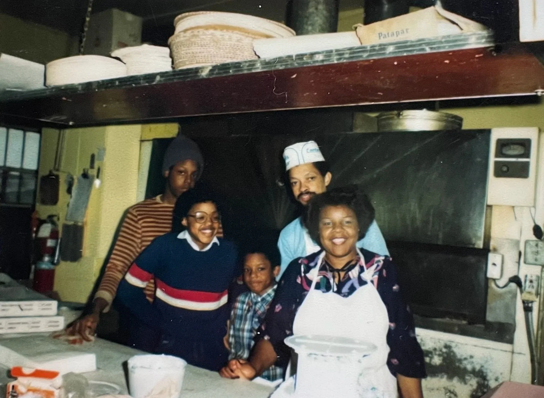 Colorful polaroid image of the Hadley family standing inside the kitchen at their bakery. Dorothy is wearing a white apron in the front, alongsdie two of her children.  Hurtis is stanidng behind Dorothy, alongside their son, wearing a striped shirt.
