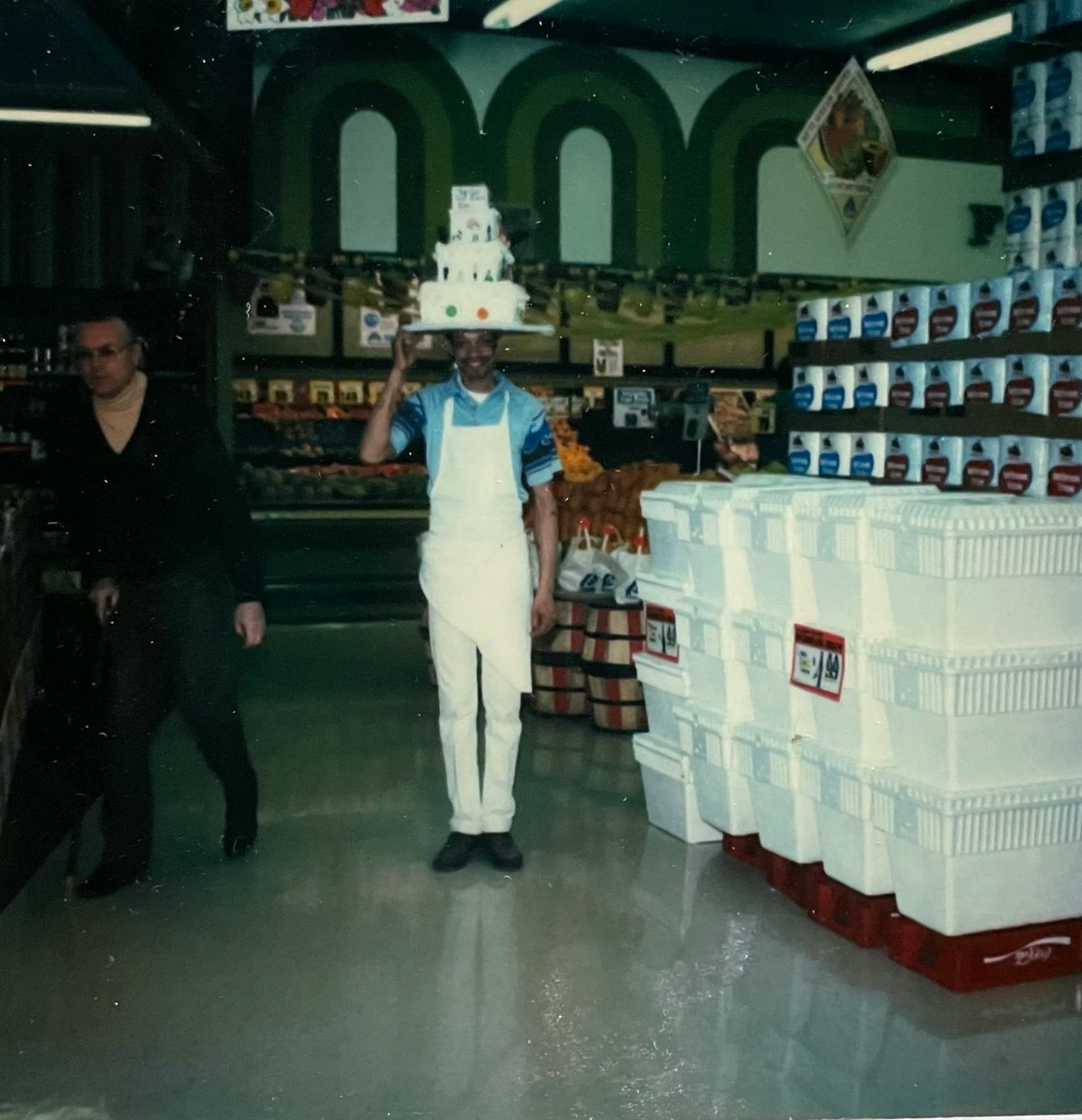 Hurtis standing in his bakery uniform. He is standing in the center of the grocery store, alongside styrofoam coolers. He is wearing a hat that looks like a three tiered cake.