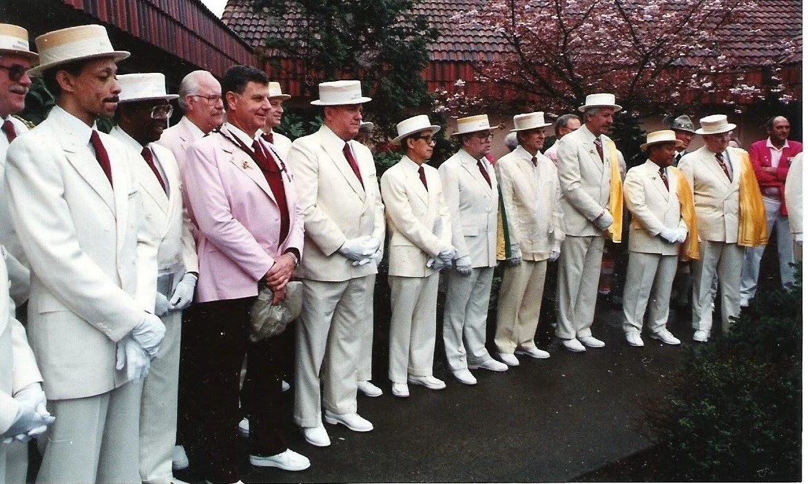 A row of men wearing white suits with white hats and red ties. Two men at the center wearing pink suit jackets. All men are standing in a row, and Hurtis Hadley is standing on the very left.