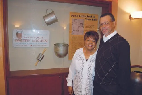 Dorothy and Hurtis Hadley standing alongside their display at OHS. Dorothy is wearing a white top. Hurtis is wearing a black vest. There is a yellow sign inside the display case, alongside a measuring cup, mixing bowl, and rolling pin. 