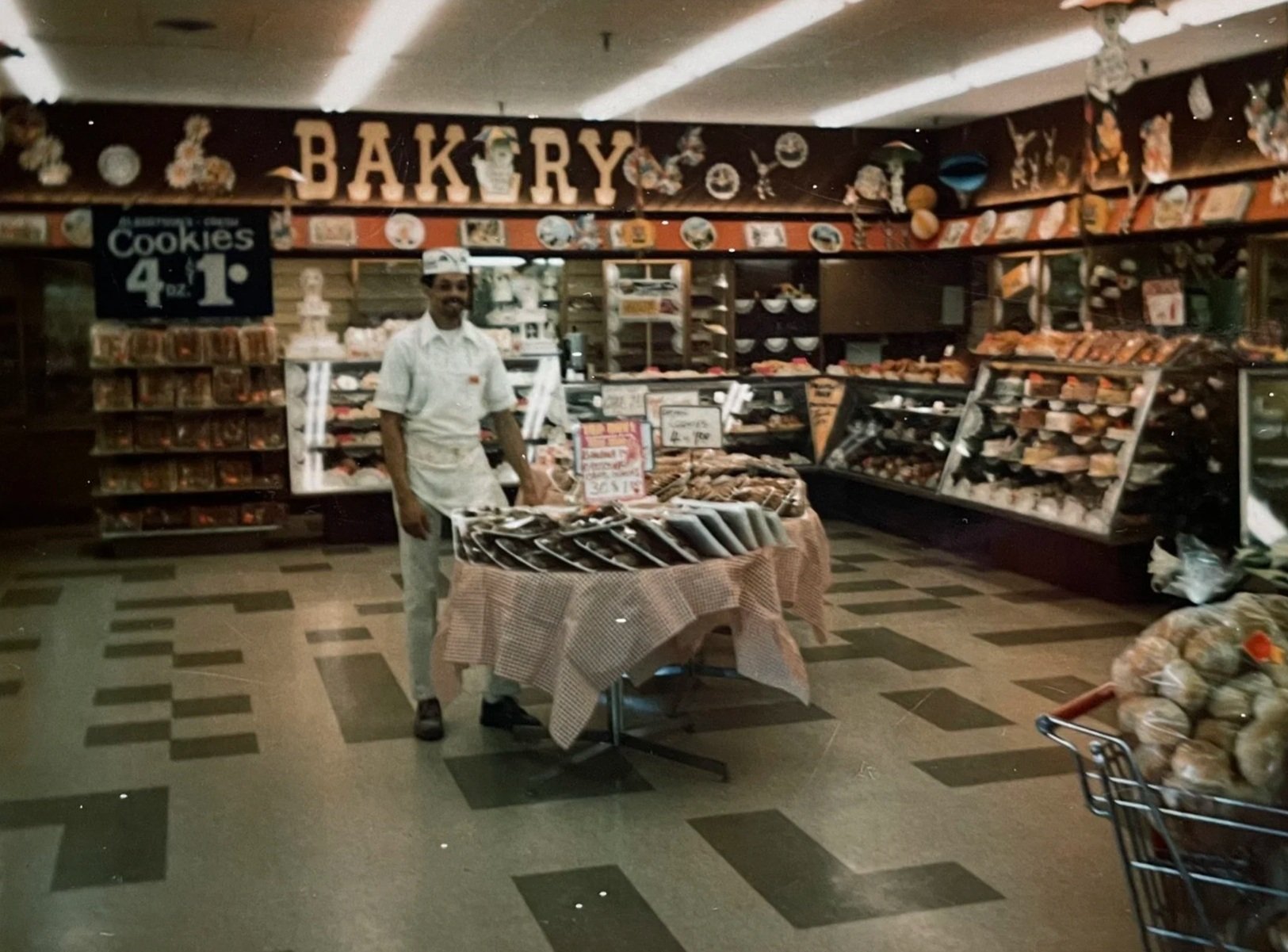 Hurtis wearing his bakers whites standing alongside a table display inside the bakery at Albertsons. There is a large "BAKERY" sign in the background, and multiple display cases filled with pastries and breads lining the walls.  