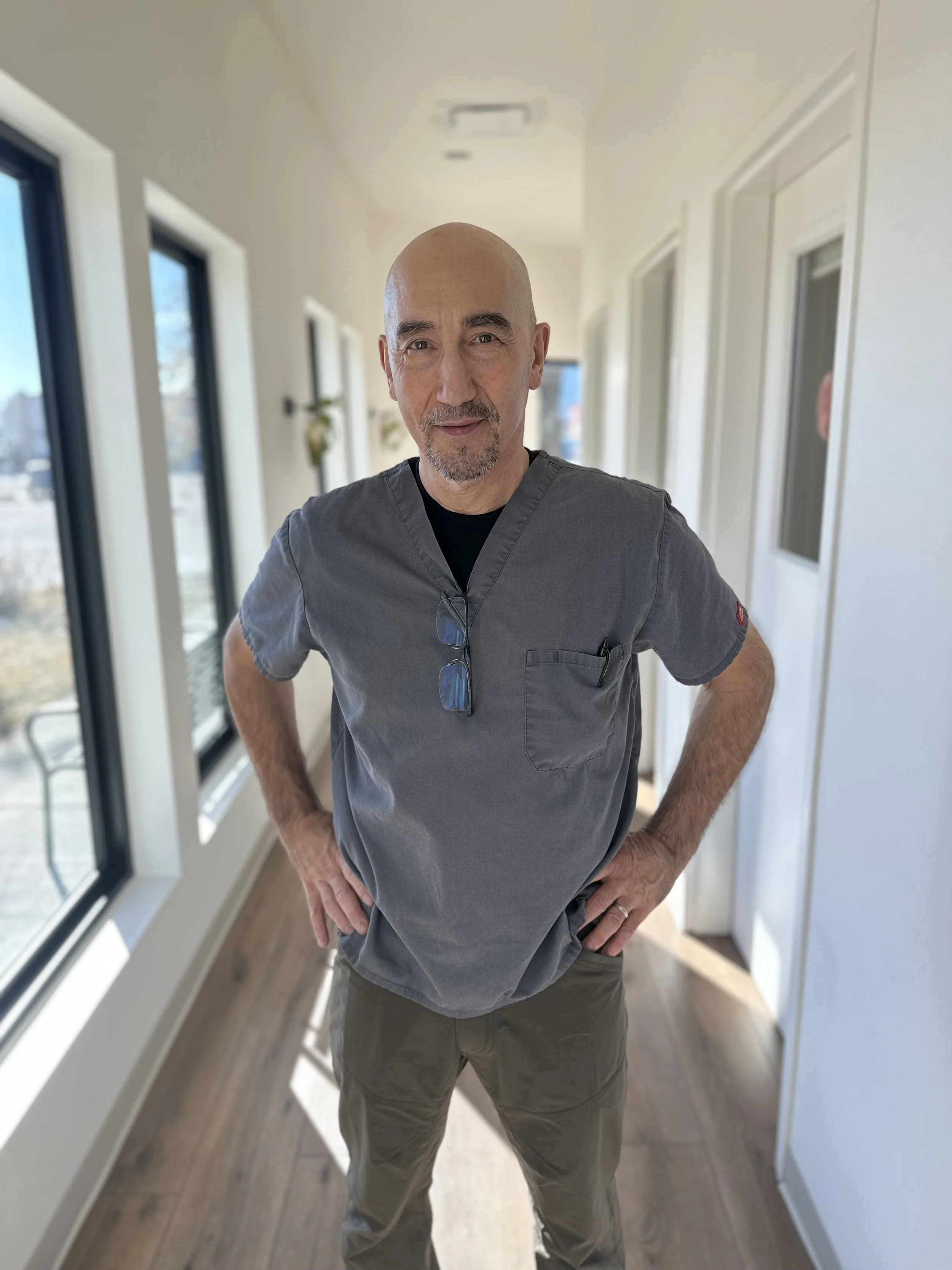 A middle-aged bald man with a beard wearing a gray medical scrub top and khaki pants is standing in a hallway with large windows, white walls, and wooden floors. He has glasses hanging from the neckline of his scrub top and is posing with his hands on his hips, smiling softly at the camera.