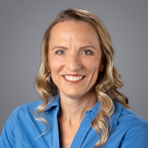 A woman with blonde hair, blue eyes, and a big smile, wearing a blue shirt, against a gray background.