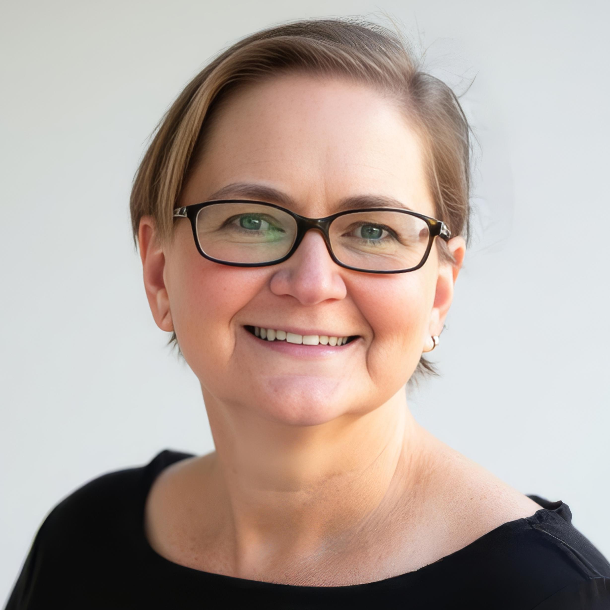 Close-up of a woman with short brown hair, wearing black-framed glasses, smiling, against a plain white background.
