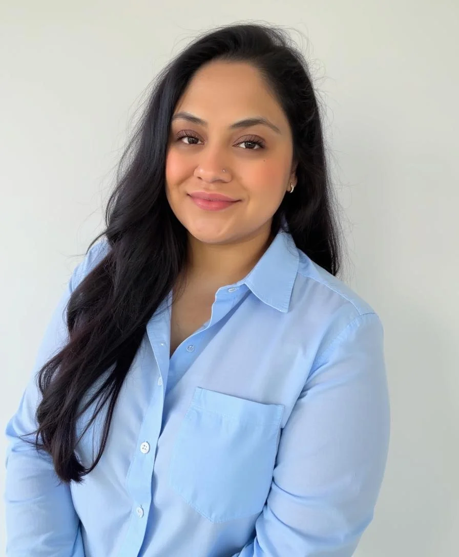 A woman with long black hair, wearing a light blue button-up shirt, smiling slightly against a plain white background.