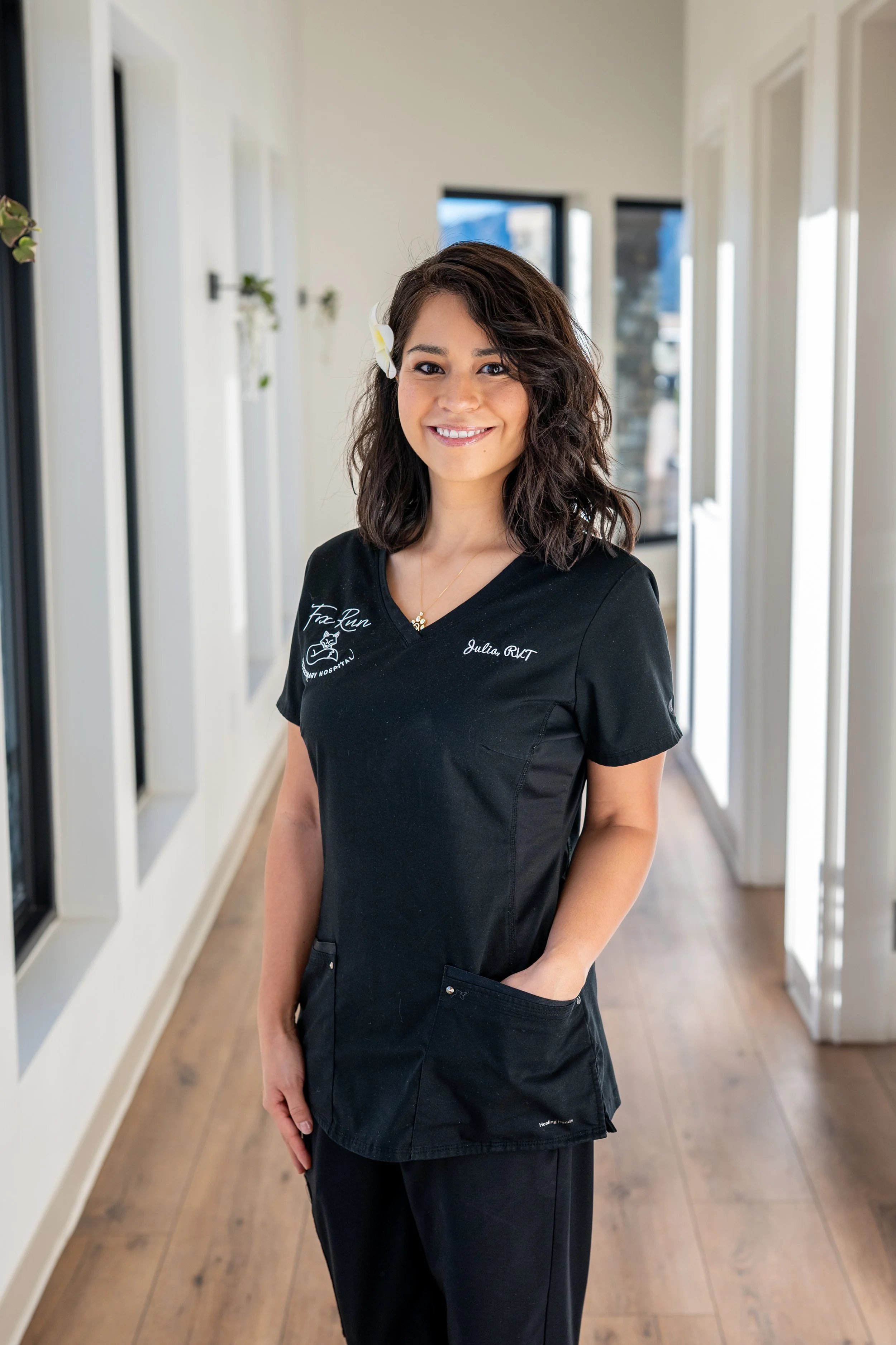 A woman wearing medical scrubs standing in a hallway with white walls and large windows, smiling at the camera.