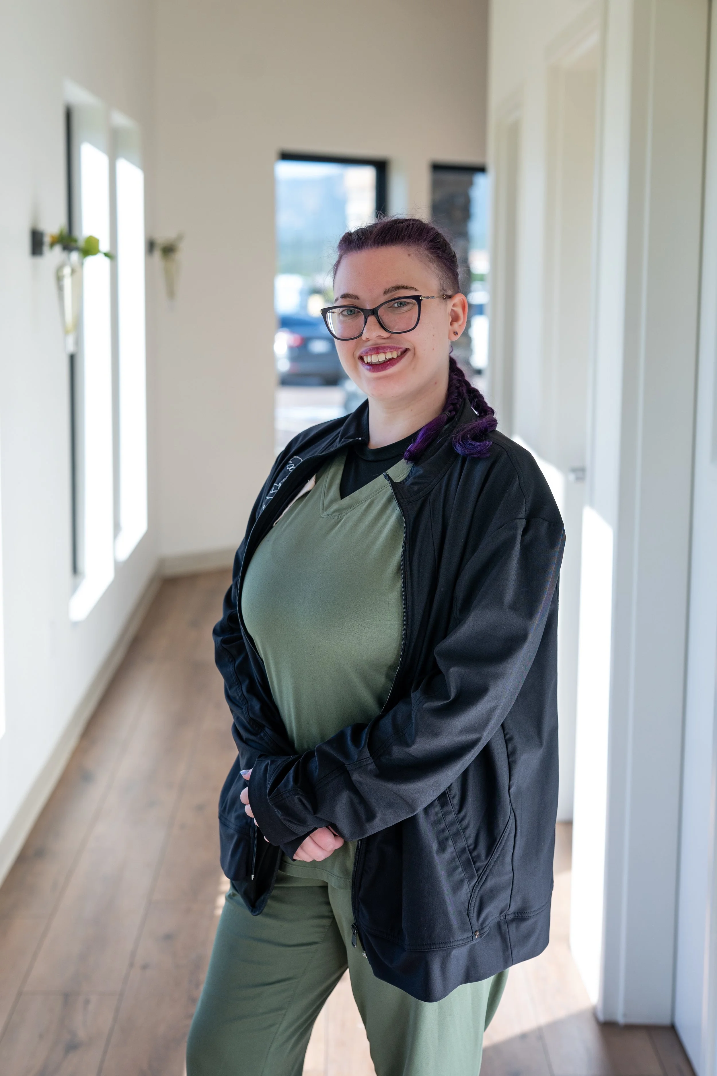 A woman with glasses and purple braided hair smiling, indoors with white walls and windows.