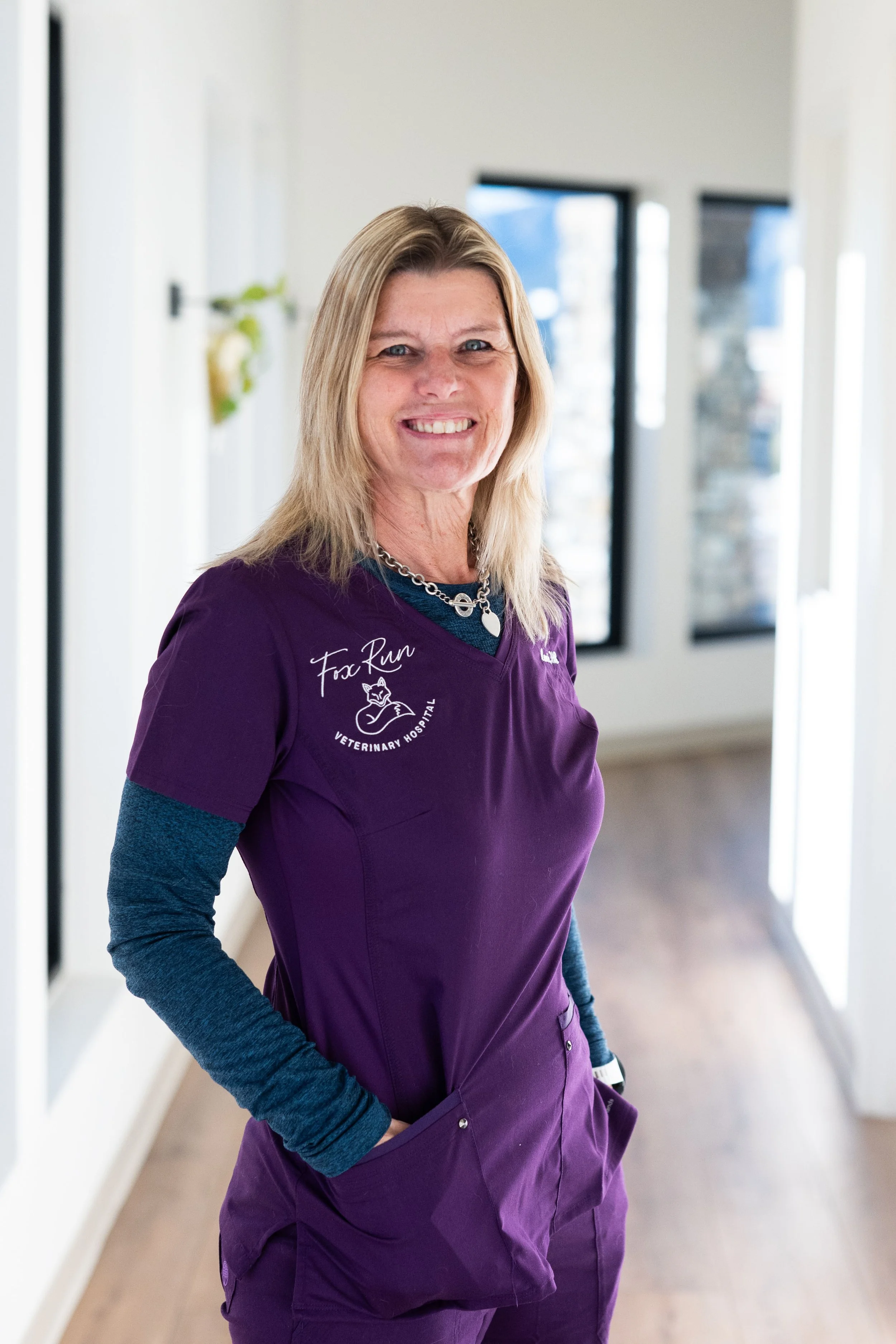 A smiling woman with blonde hair wearing purple veterinary scrubs stands in a bright indoor setting with windows in the background.