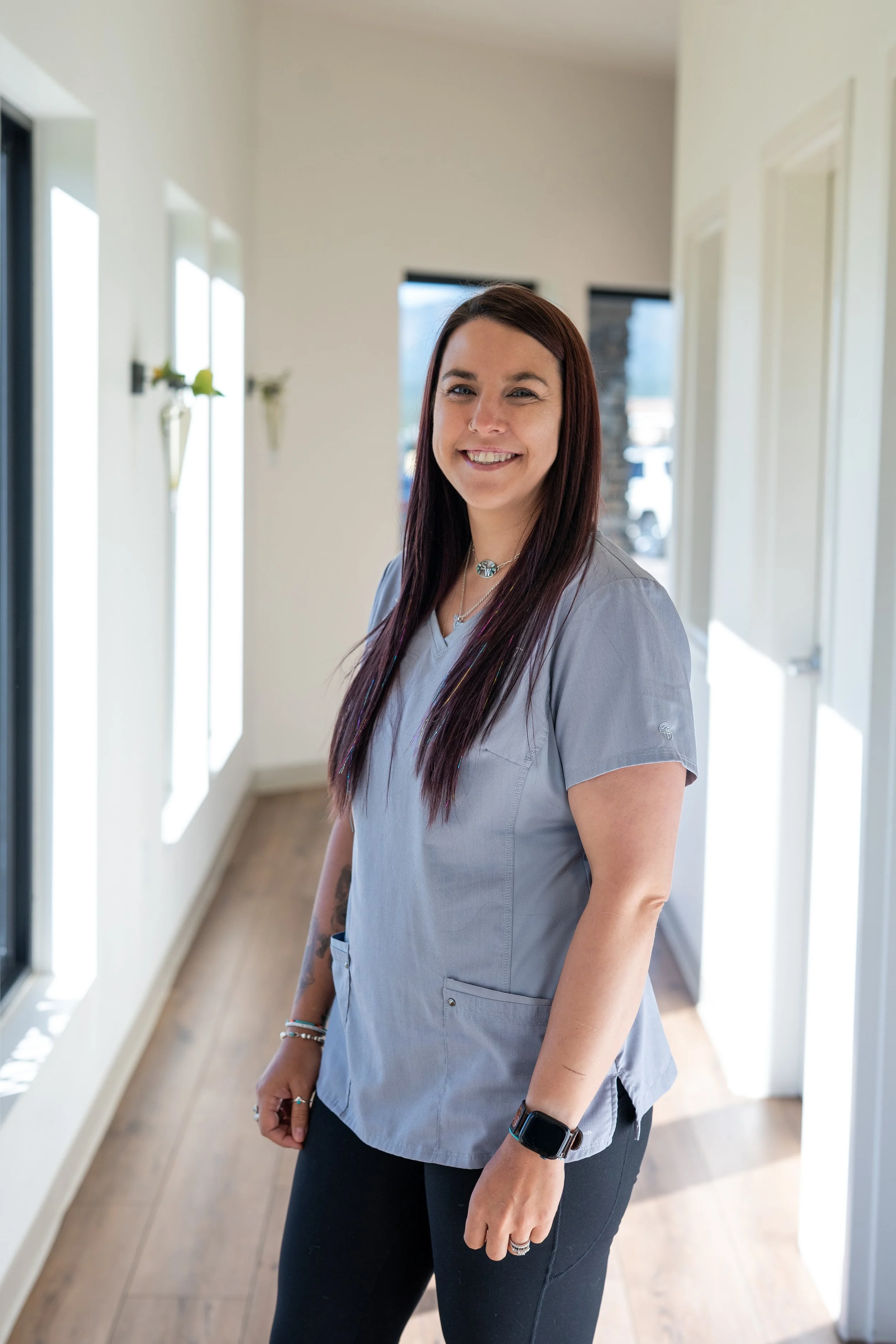 A woman with long dark red hair smiling in a brightly lit hallway with white walls and wooden flooring.