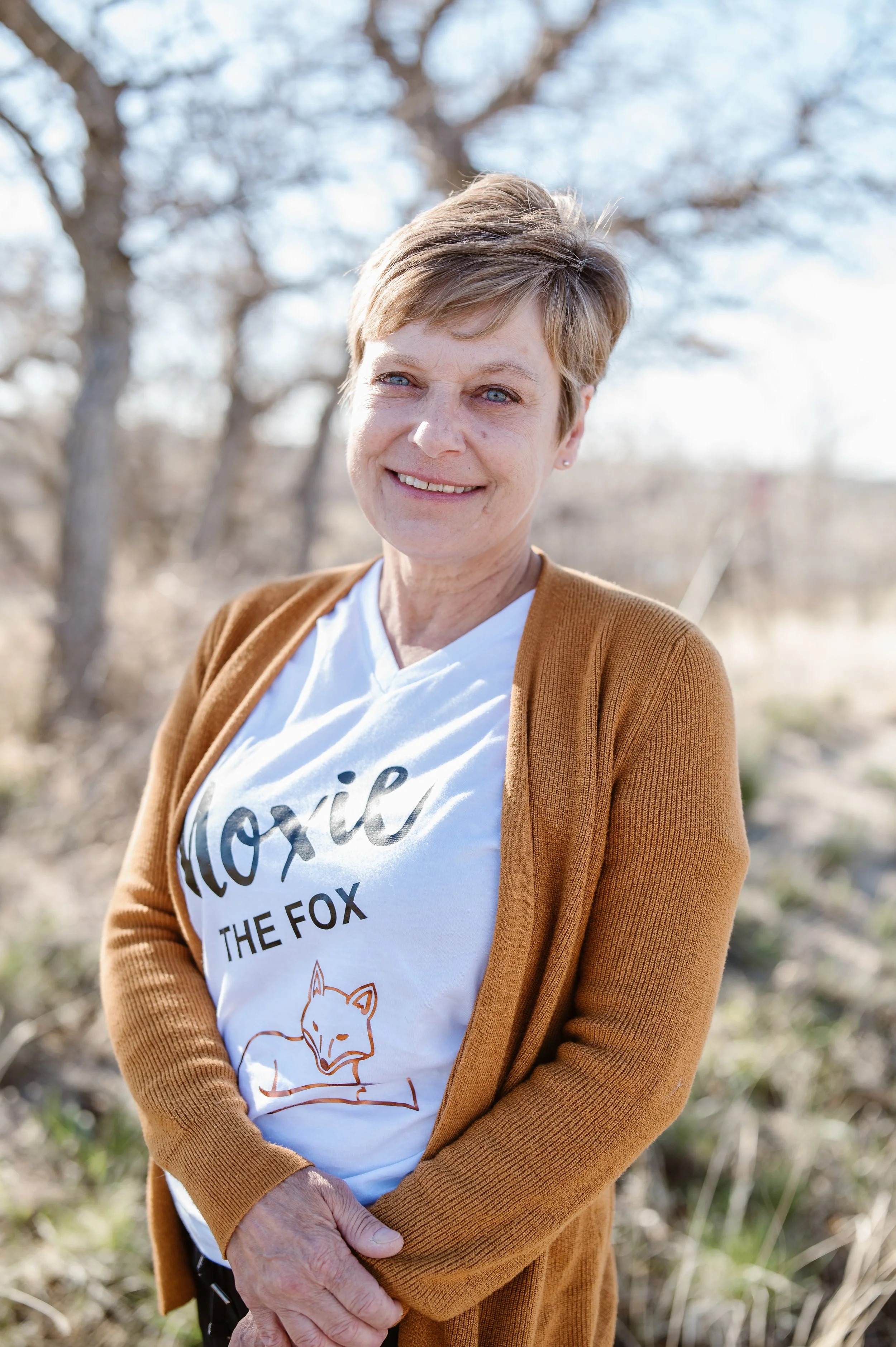 A smiling woman with short hair standing outdoors in front of trees and bright blue sky, wearing a white T-shirt with a fox illustration and text, and a brown cardigan.