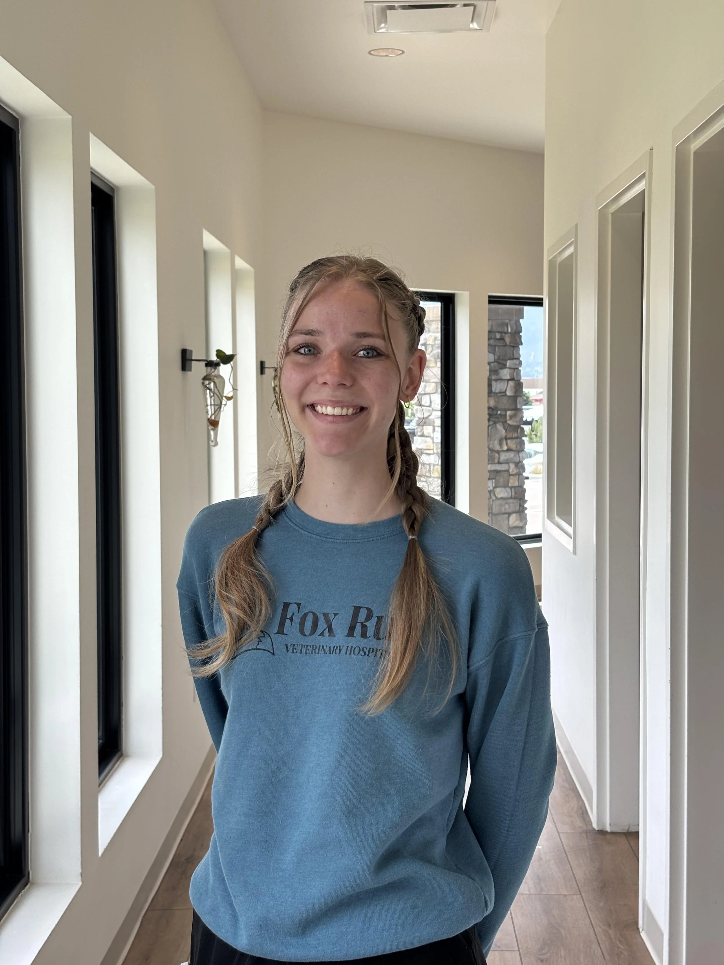 A young woman with braided hair, blue eyes, and freckles, smiling while standing indoors near a window. She is wearing a blue long-sleeve shirt with text that reads 'Fox Run Veterinary Hospital.'
