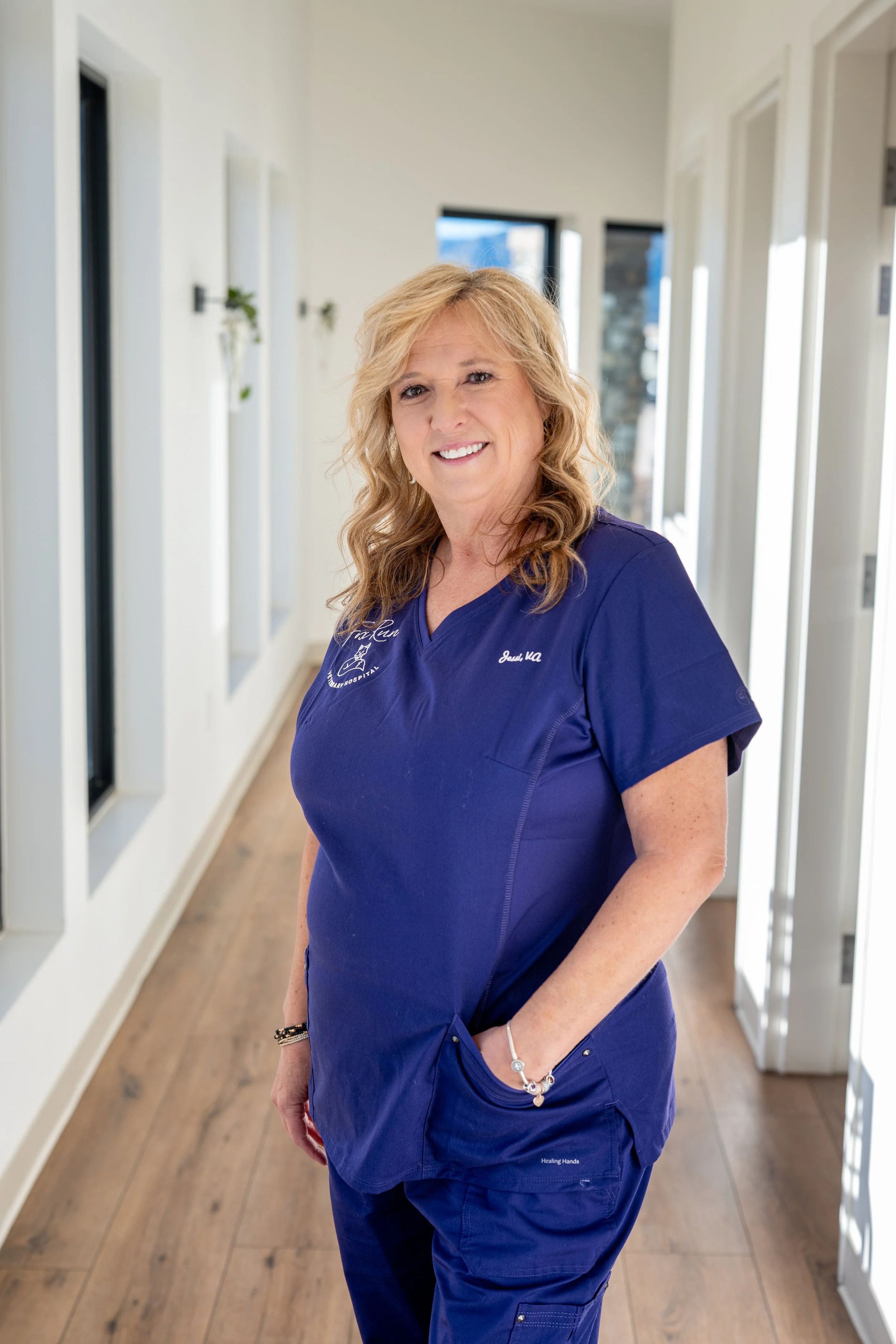 A smiling woman in blue medical scrubs standing in a bright hallway with large windows and wooden floors.