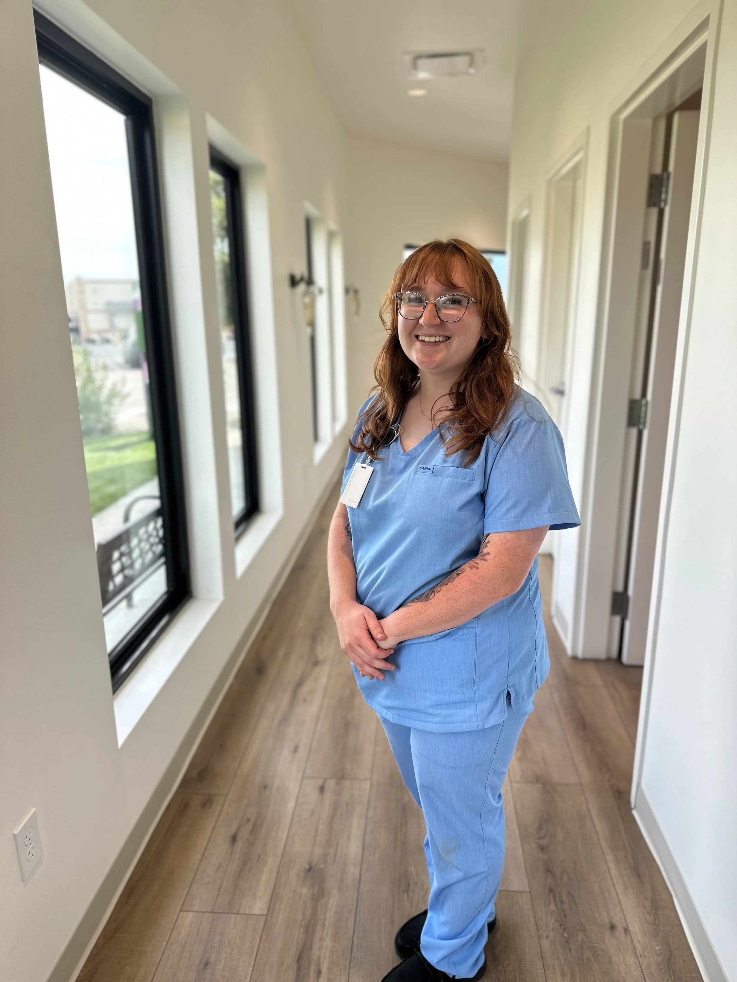 A smiling woman with red hair and glasses in blue medical scrubs standing in a hallway near a window with black trim.