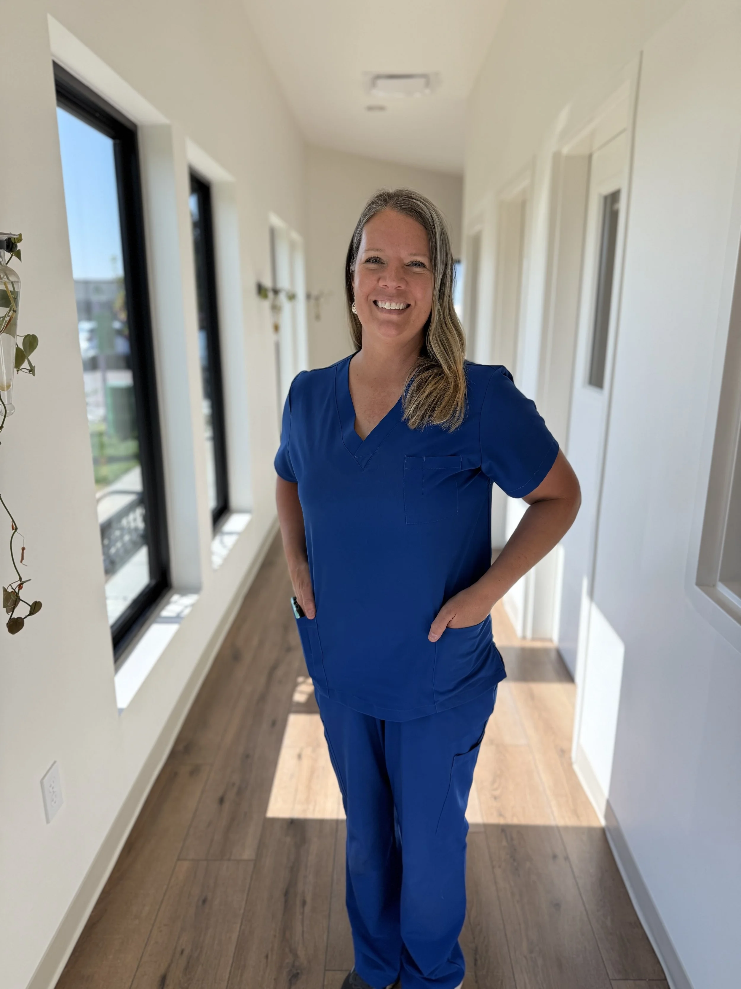 A woman in blue medical scrubs standing in a hallway with white walls, wooden flooring, and large windows, smiling at the camera.
