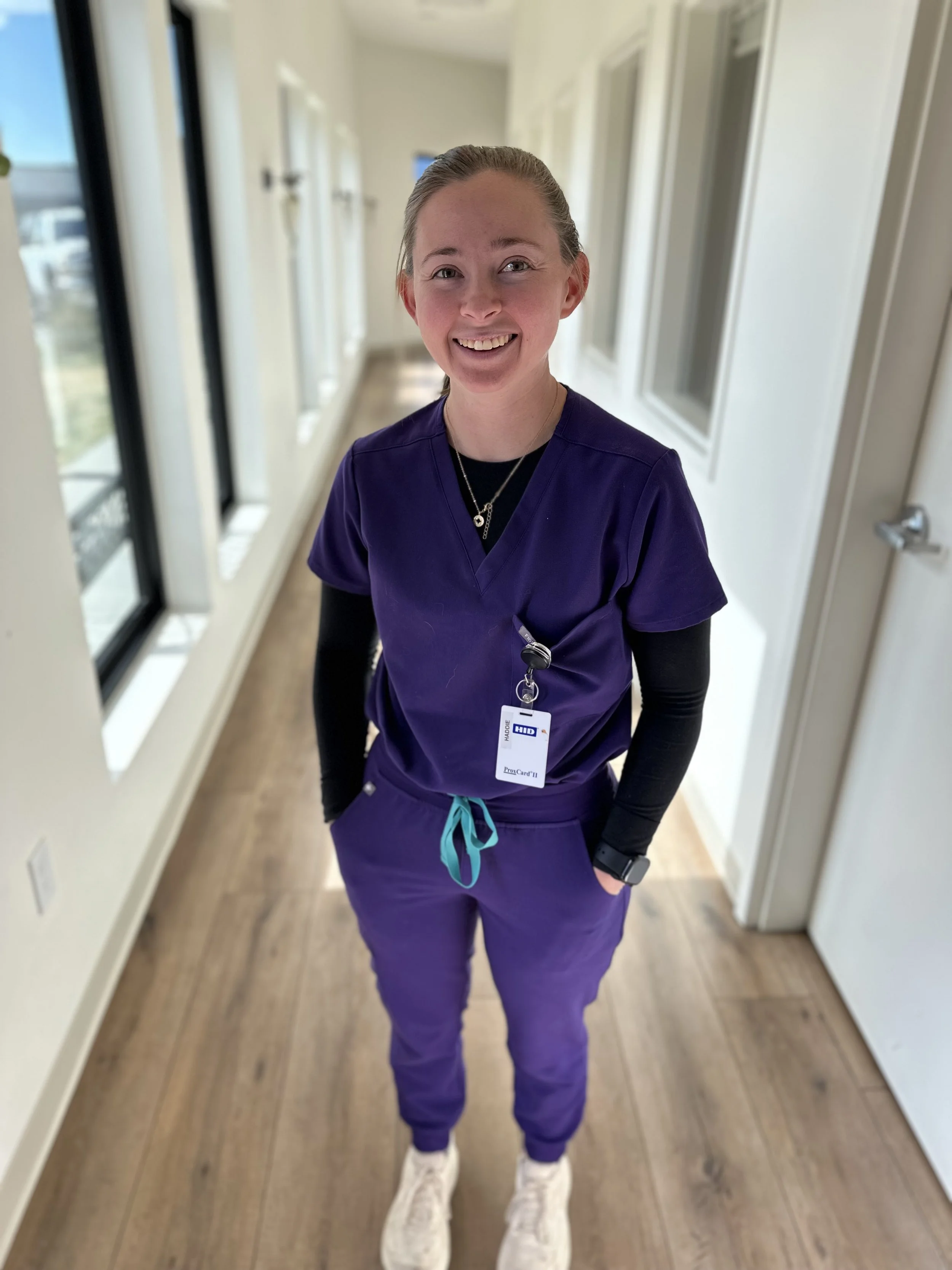 A woman in purple scrubs standing inside a brightly lit hallway with large windows on one side, smiling at the camera.