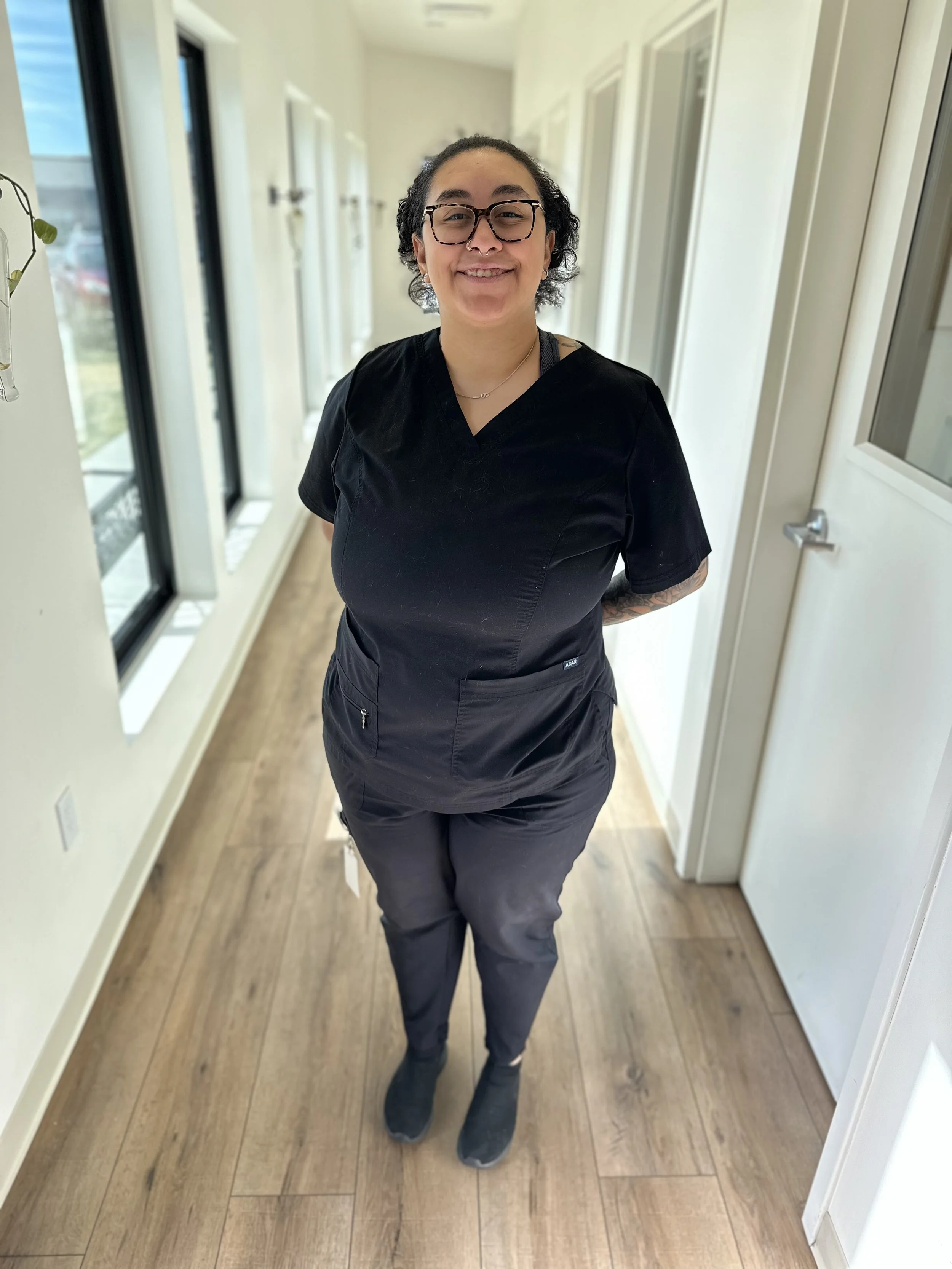 A person with curly hair, glasses, and a septum piercing, standing in a hallway with light-colored walls and wooden flooring. They are smiling and wearing black scrubs and sneakers.