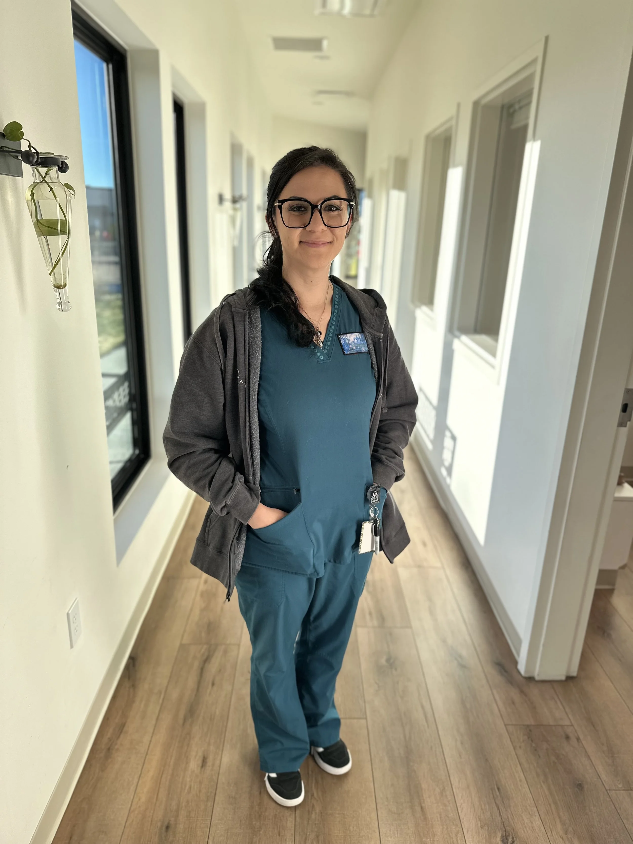 A woman in teal medical scrubs and a gray hoodie with ID badge stands in a bright hallway with wooden flooring, windows, and wall-mounted lights.