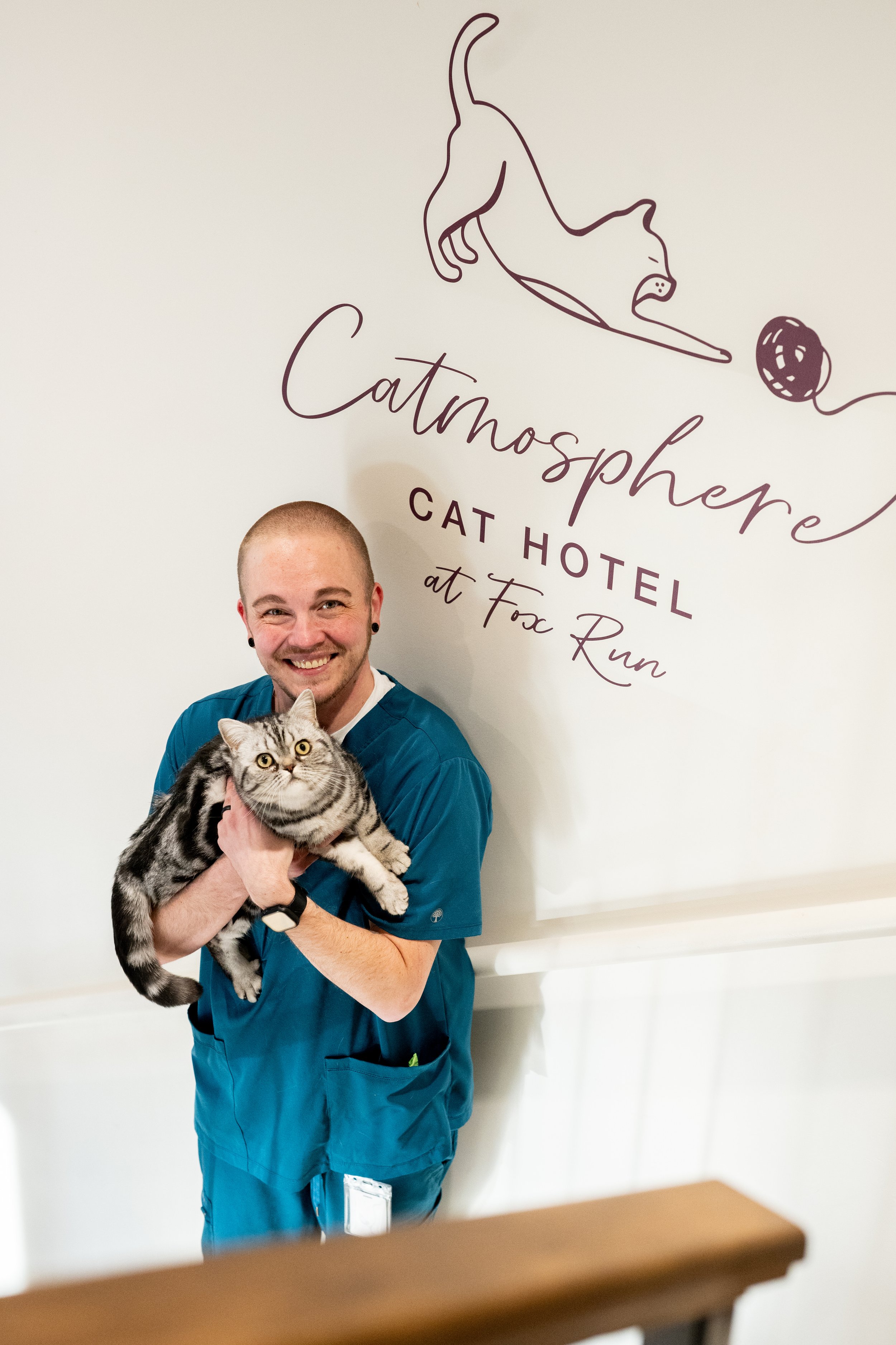 A man in scrubs holding a gray tabby cat at a veterinary clinic or animal hospital. Behind them, a sign reads 'Catomosphere Cat Hotel at Fox Run' with a line drawing of a lying cat and a ball of yarn.