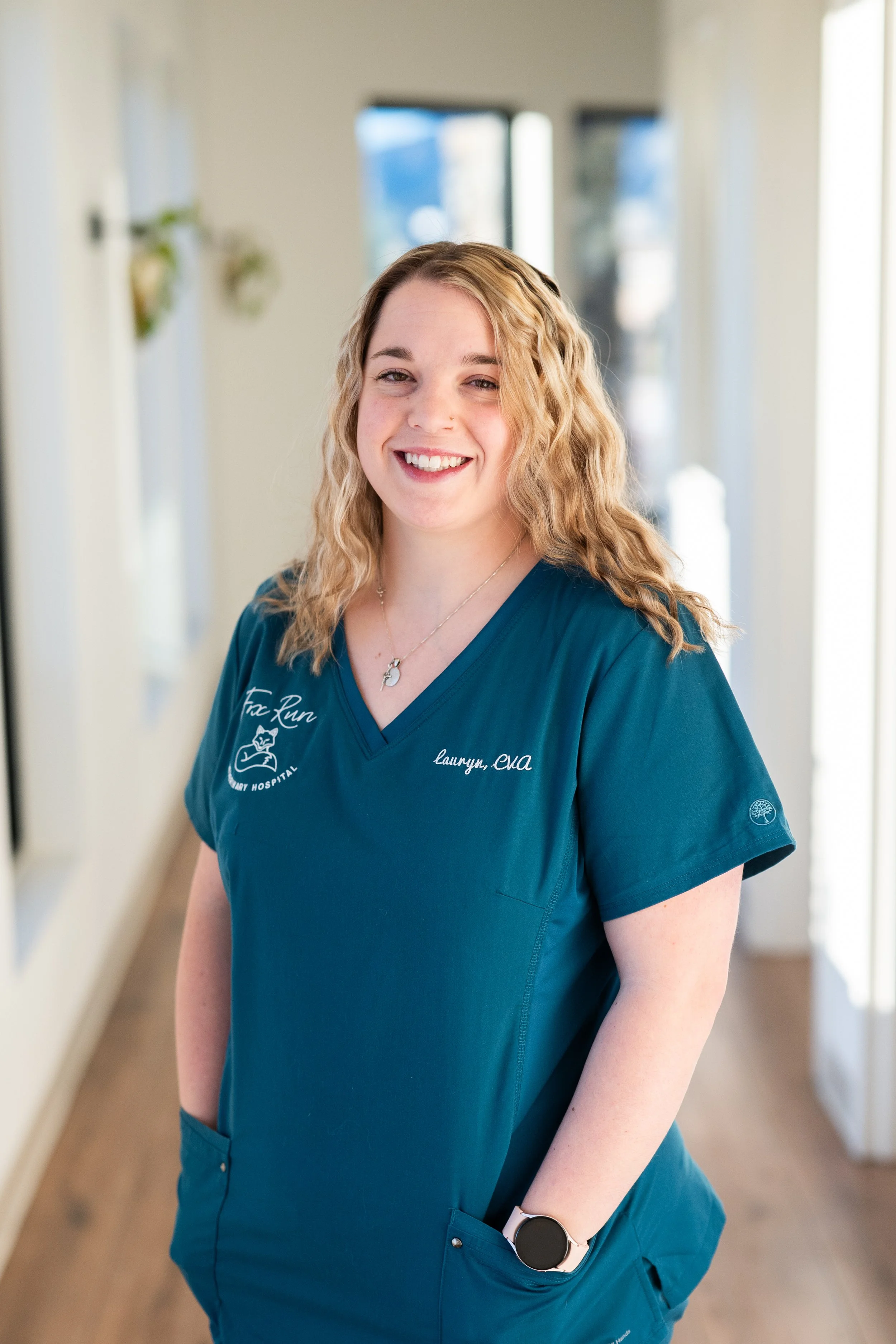 A smiling young woman with blonde, curly hair standing in a hospital corridor wearing teal scrubs with the name tag 'Lauryn, CNA' and a smartwatch on her left wrist.