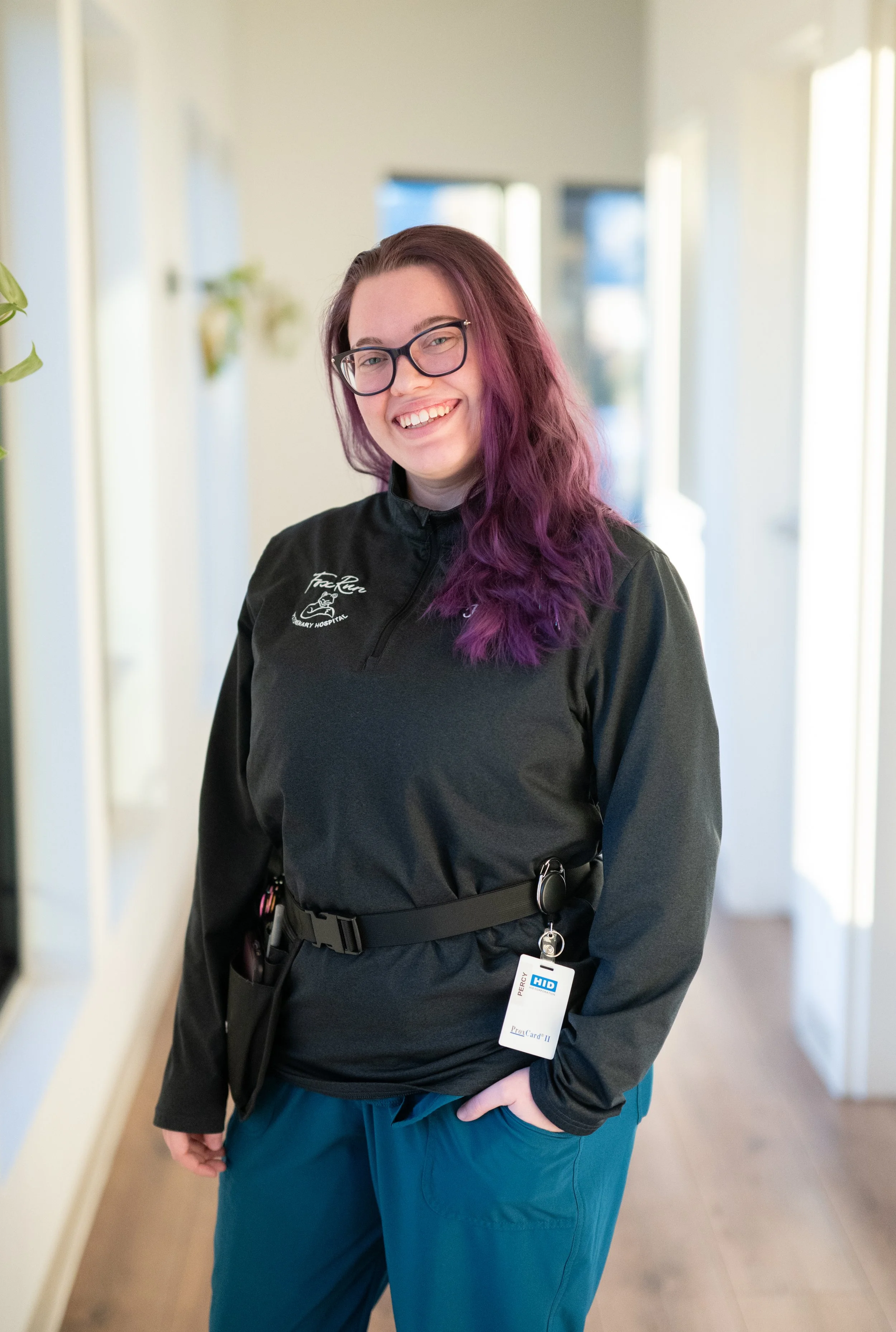 Person with purple hair wearing glasses and a black shirt, standing in a hallway.