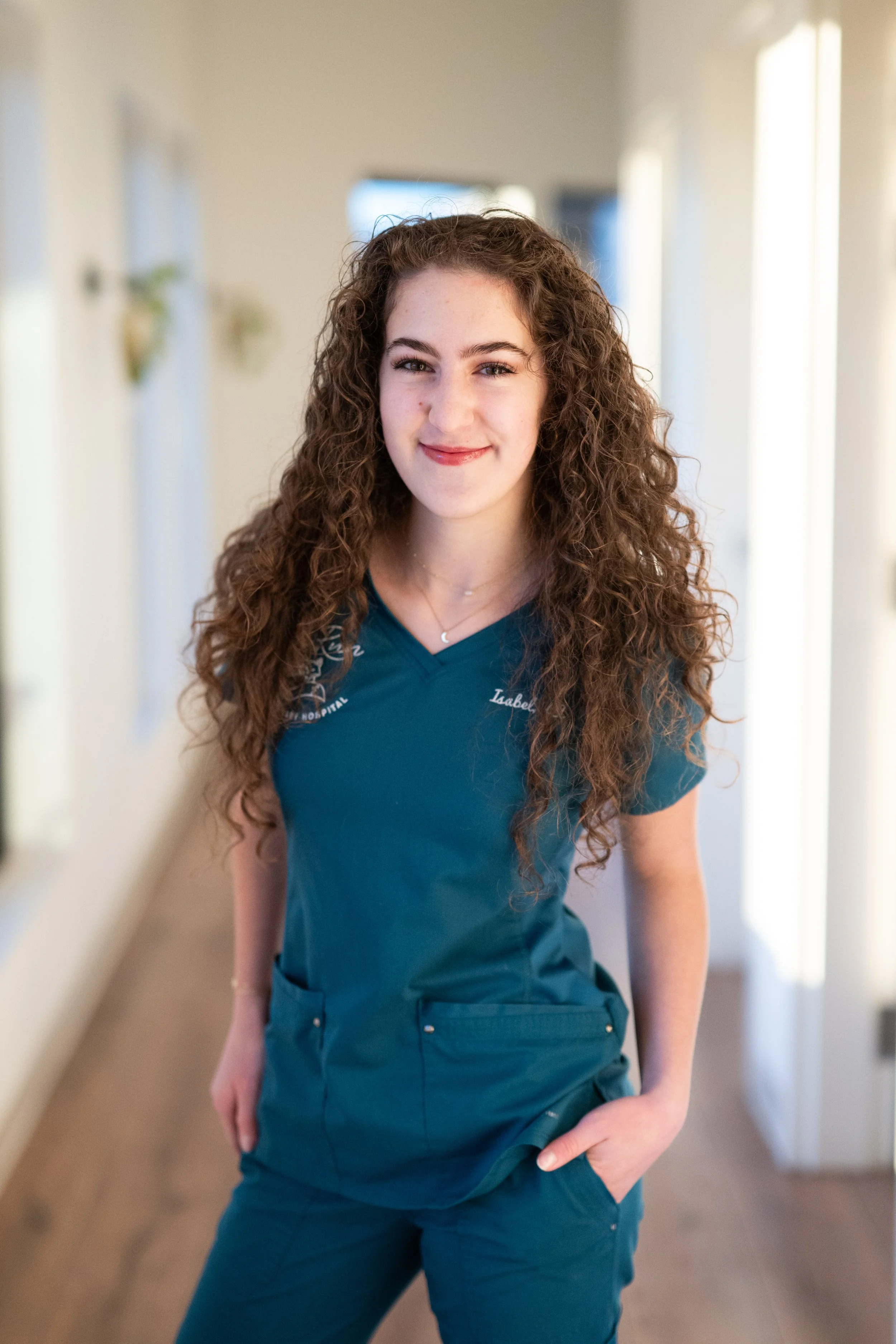 A young woman with long, curly brown hair and fair skin in a hallway, wearing blue medical scrubs with her left hand in her pocket, smiling at the camera.