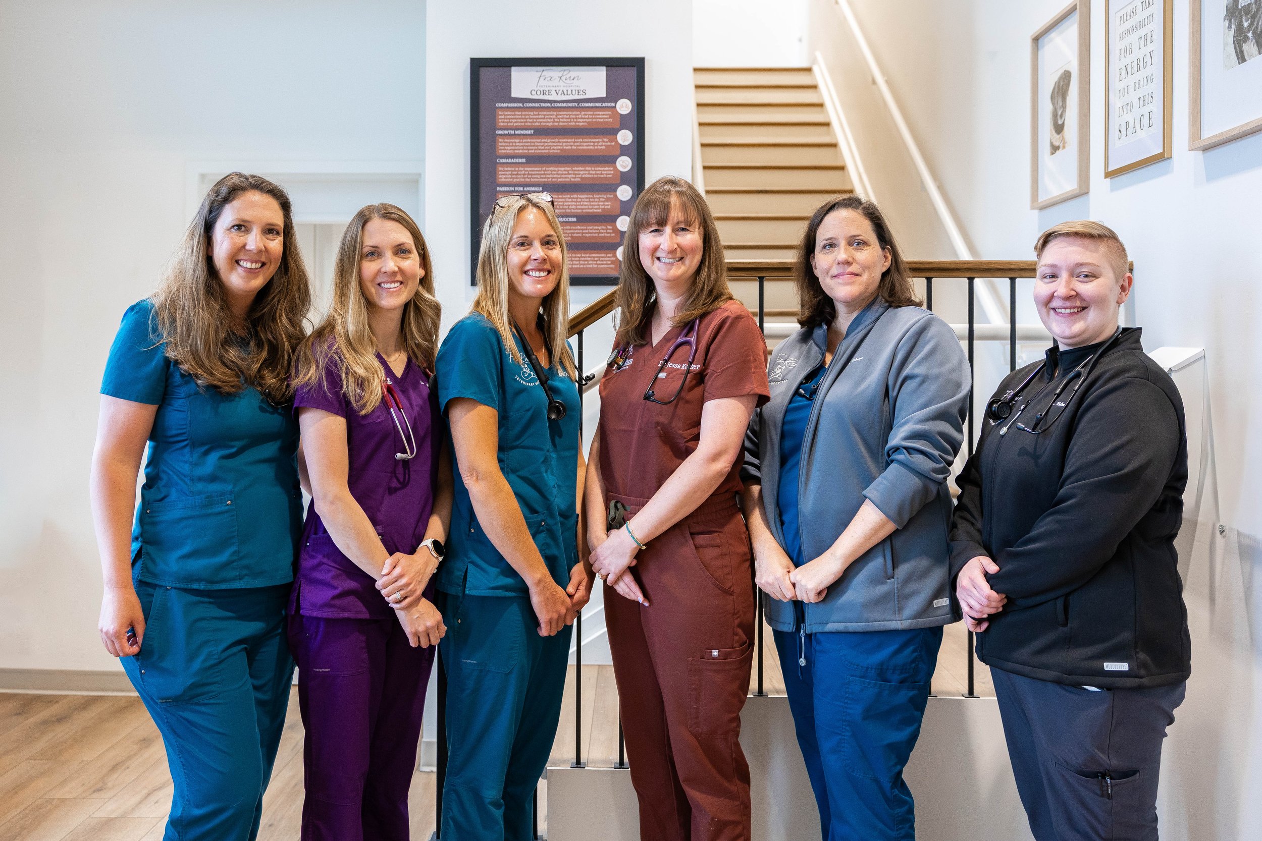 Group of six female healthcare professionals standing together indoors, smiling, wearing scrubs and lab coats with stethoscopes around their necks.