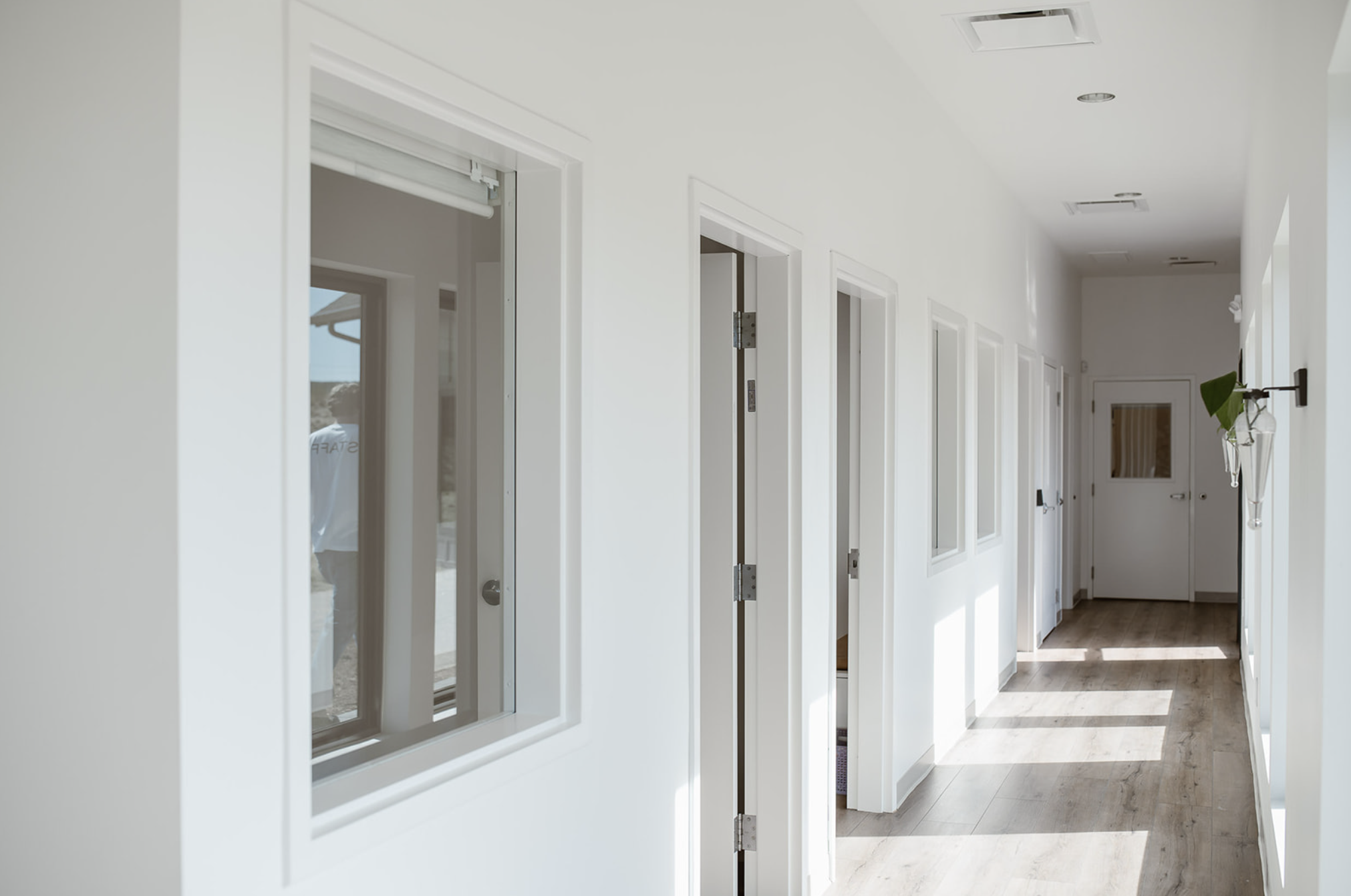 A bright hallway with white walls and multiple doors, some opening to rooms, with wood flooring and natural light streaming in.