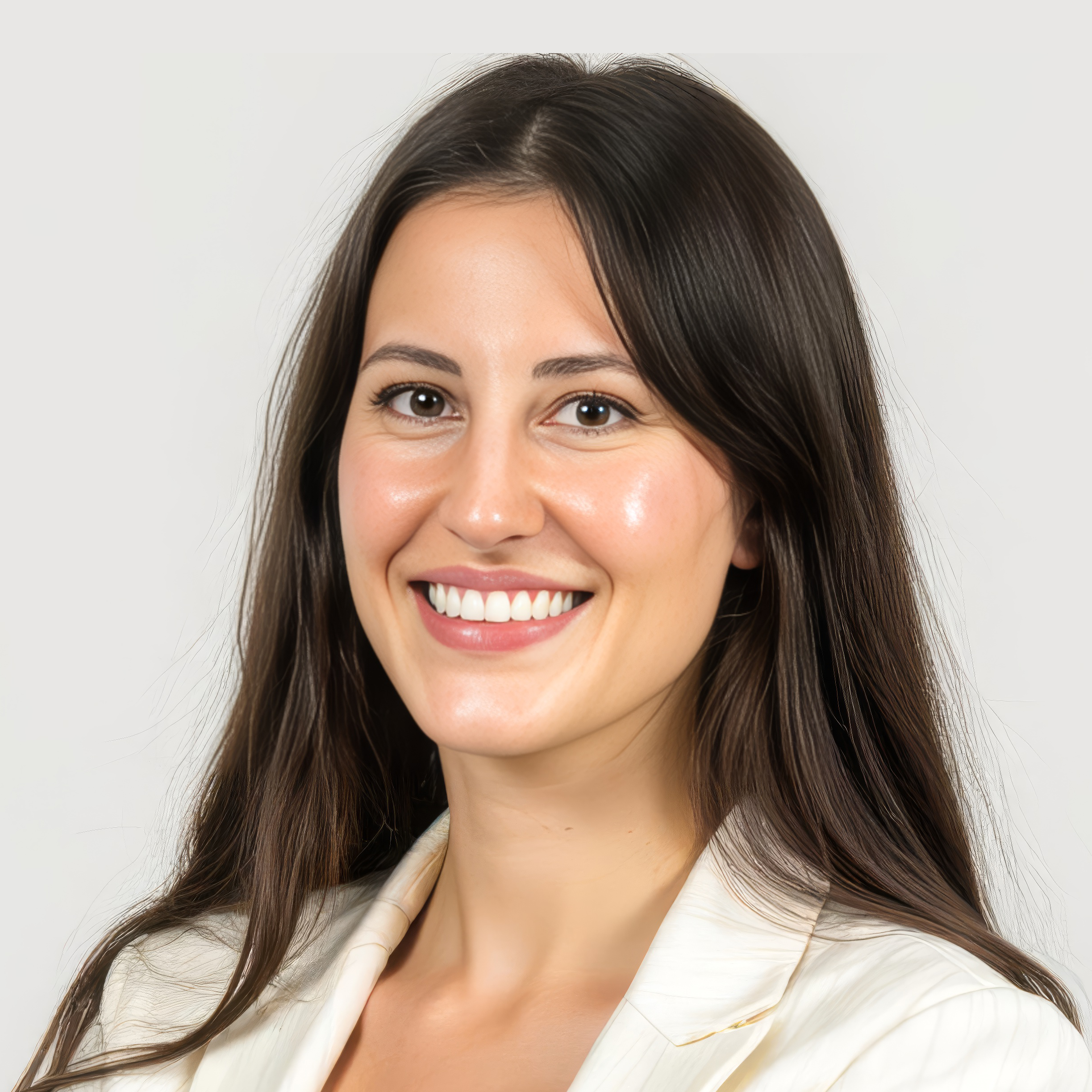 Close-up of a young woman with long brown hair smiling and touching her chin with her hand, wearing a white blazer, with a light background.