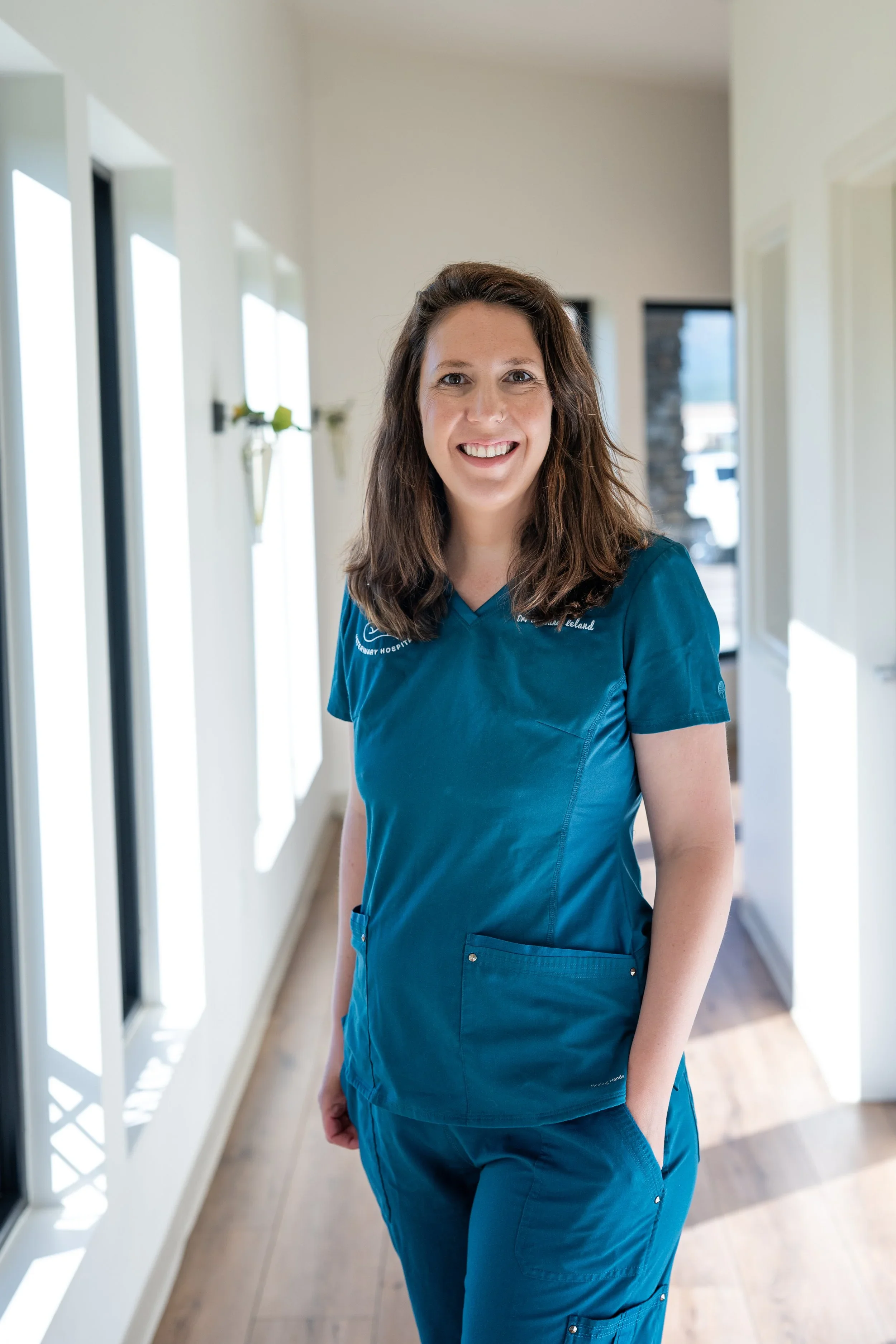 A woman with brown hair wearing blue scrubs standing in a brightly lit hallway with white walls and large windows.