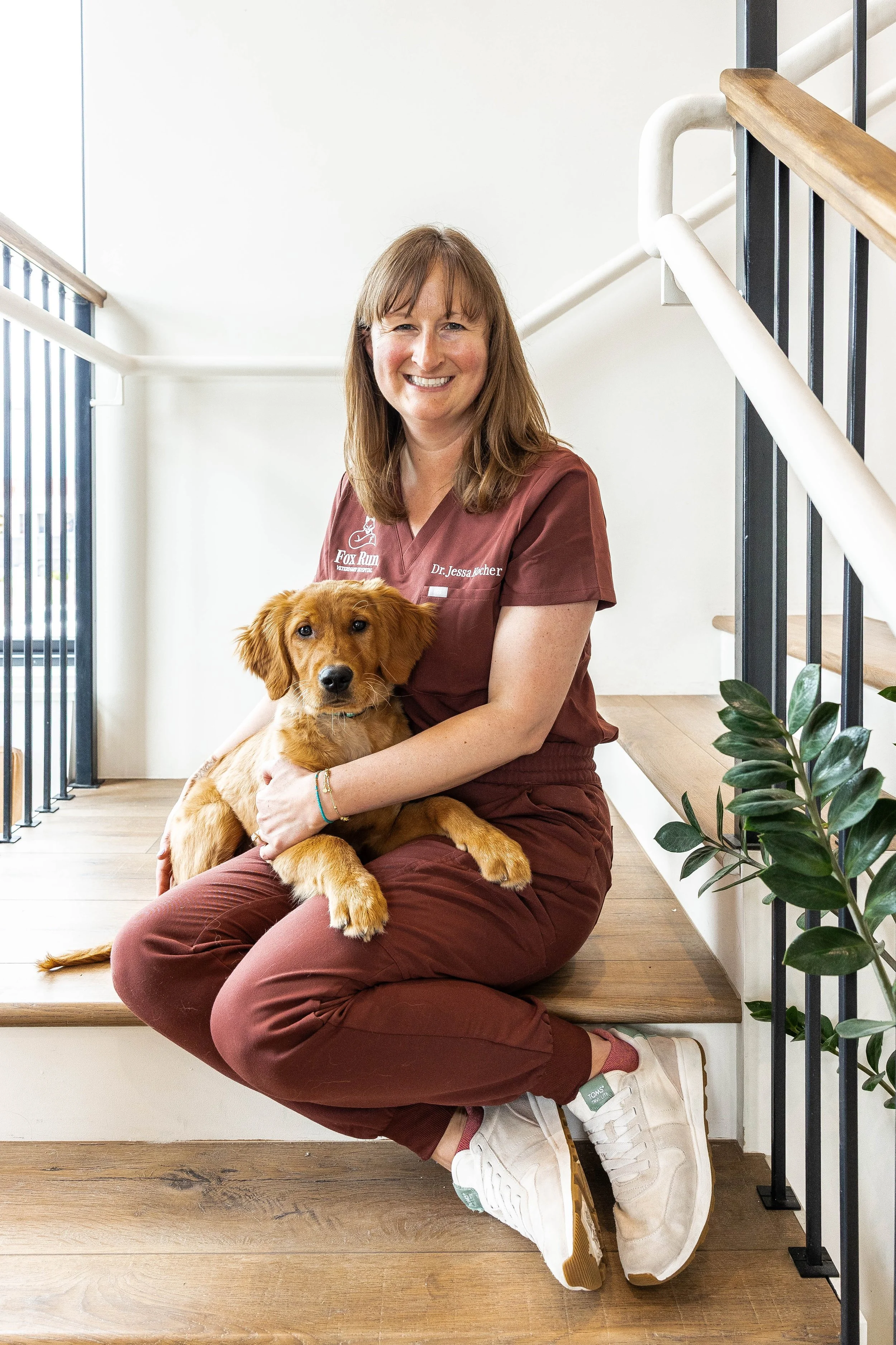 A woman with shoulder-length brown hair, wearing a nurses uniform with her name tag, is sitting on stairs and holding a Golden Retriever puppy. The background shows a white wall and a staircase with black railing and wooden handrail.