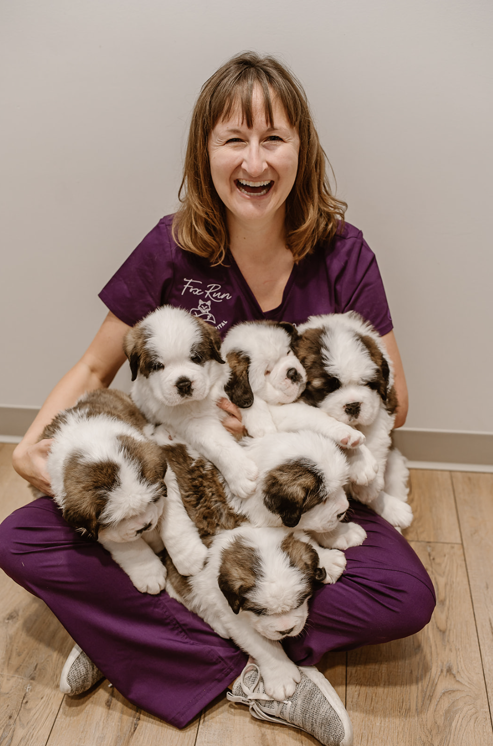 A woman in purple scrubs sitting on the floor, smiling, while holding several adorable black and white and brown and white puppies on her lap and around her.