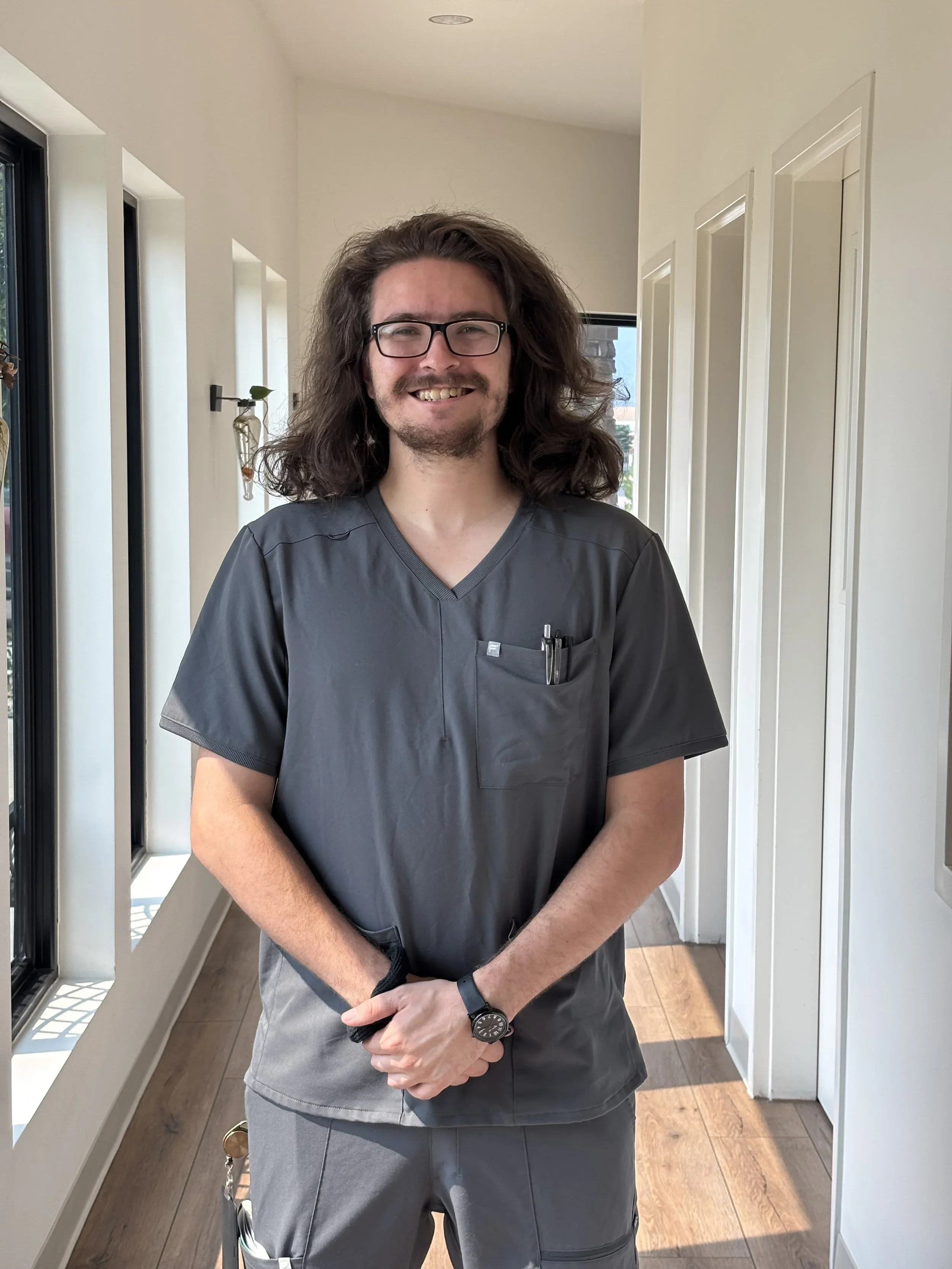 A man with long curly hair, glasses, and a beard, wearing gray medical scrubs, stands inside a well-lit house near windows with a sunny view outside, smiling at the camera.