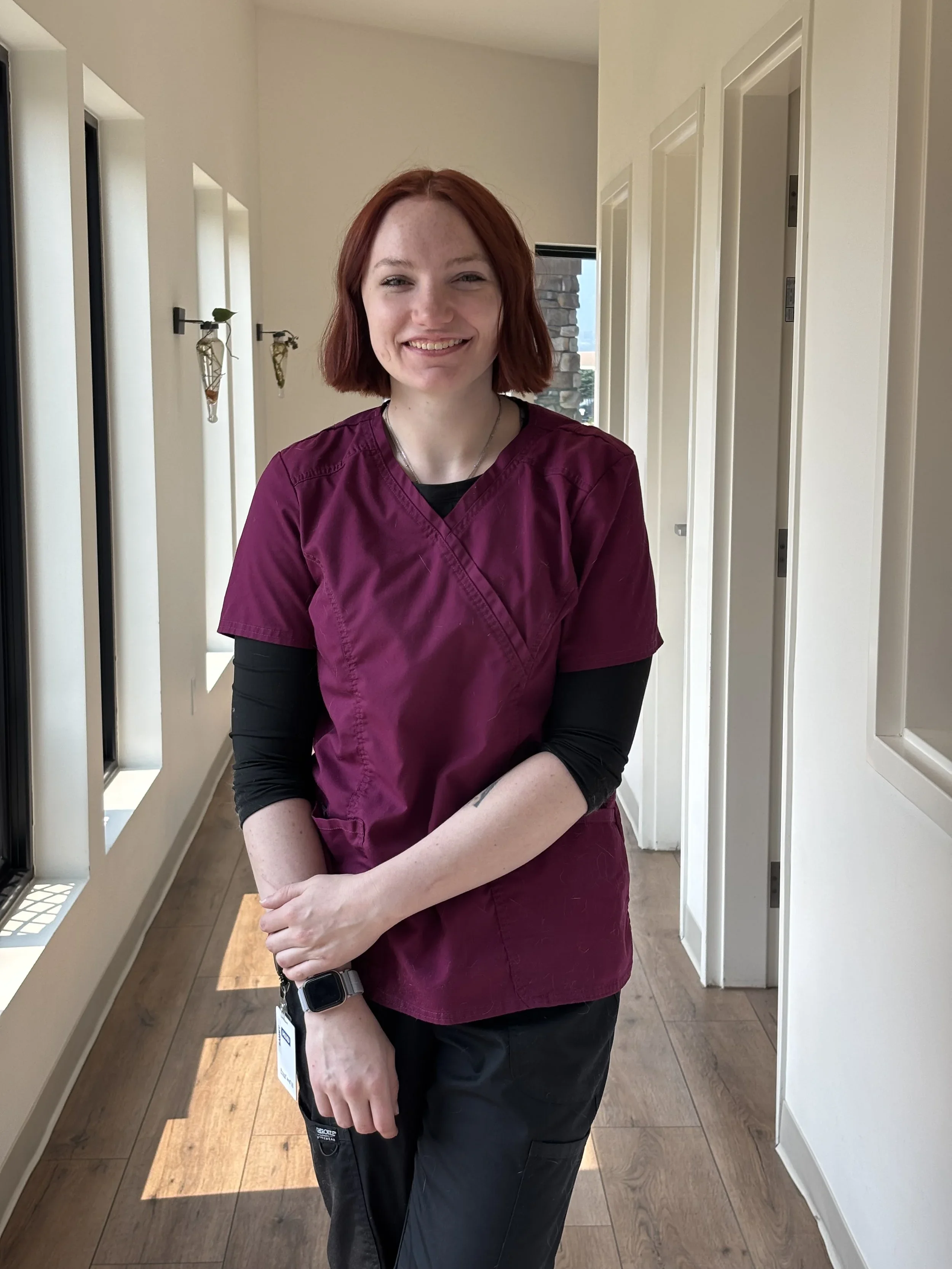 A woman with red hair and freckles wearing a maroon medical scrub top and black pants, standing in a hallway with white walls and wooden flooring, smiling at the camera.