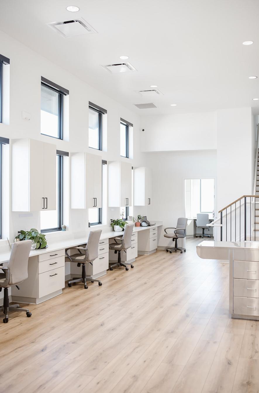 An empty modern office with white walls, multiple windows, beige office chairs, white desks, and wooden flooring.
