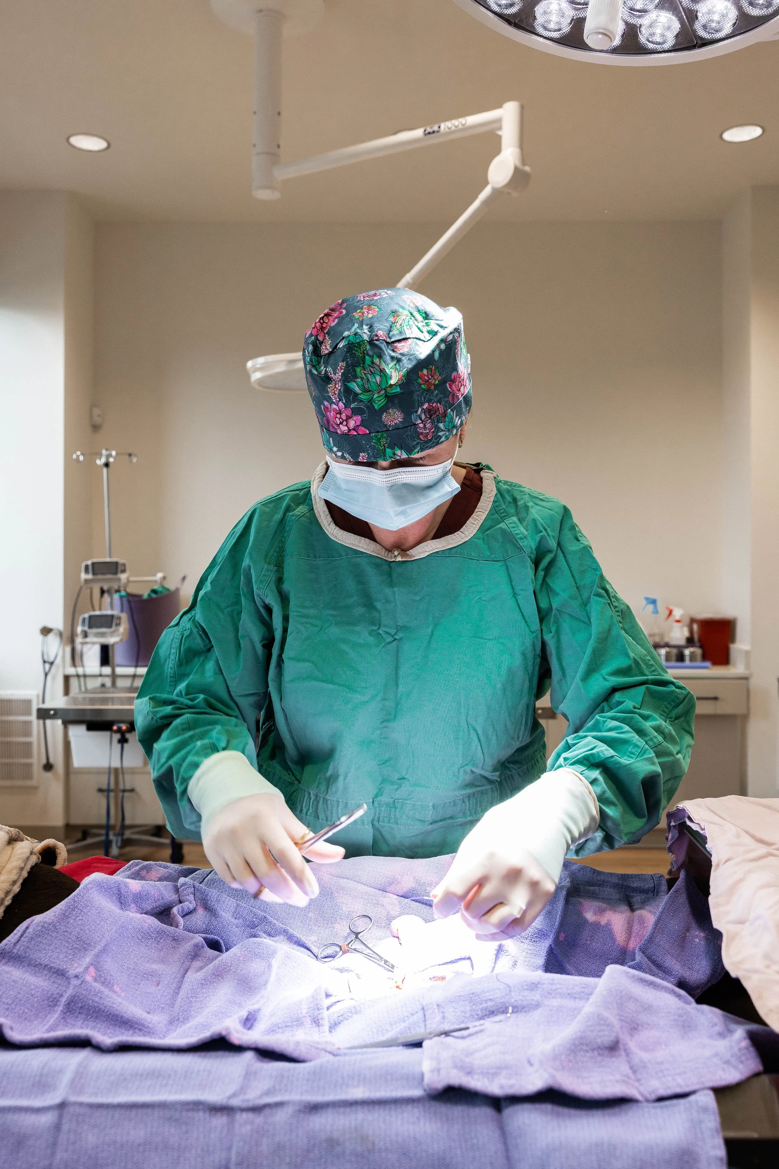 A healthcare professional wearing a floral surgical cap, face mask, green surgical gown, and gloves performing surgery in an operating room, with medical equipment visible in the background.