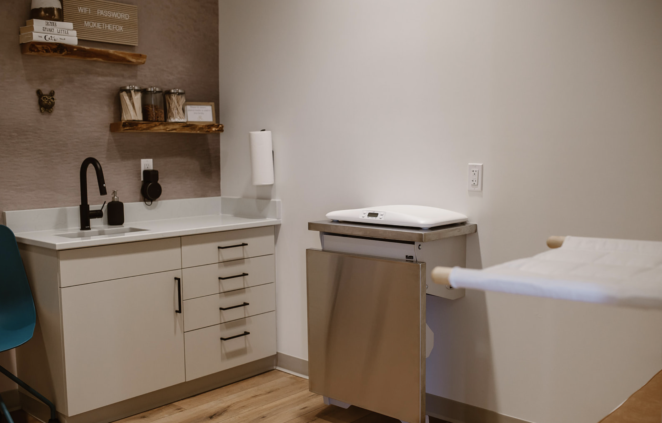 Medical examination room with a white countertop, black sink faucet, paper towel holder, and wooden shelves with containers and books, next to a bed with white linens and wooden side rails.