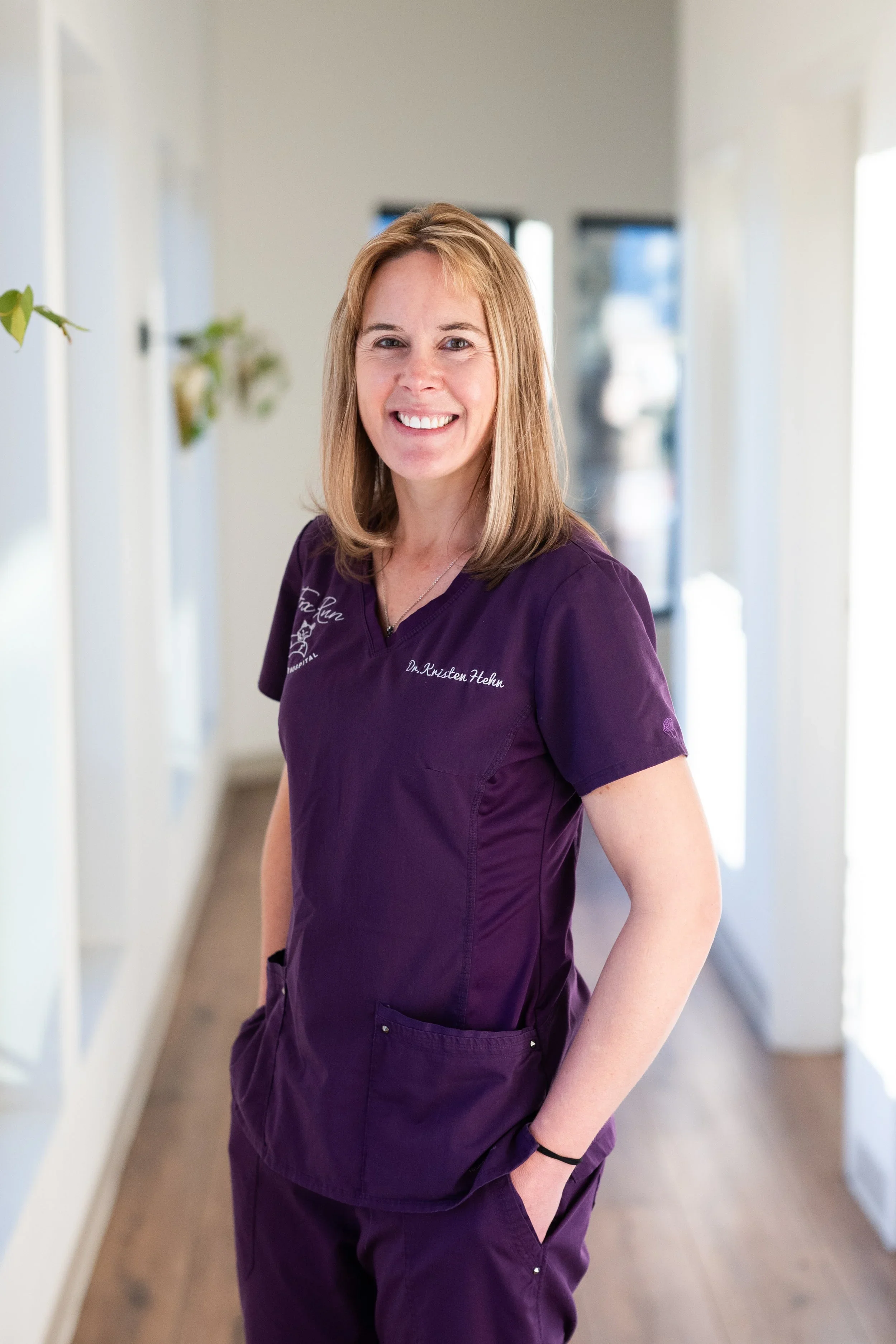 A smiling woman with shoulder-length blond hair wearing a purple medical uniform standing in a hallway with white walls and wooden flooring.